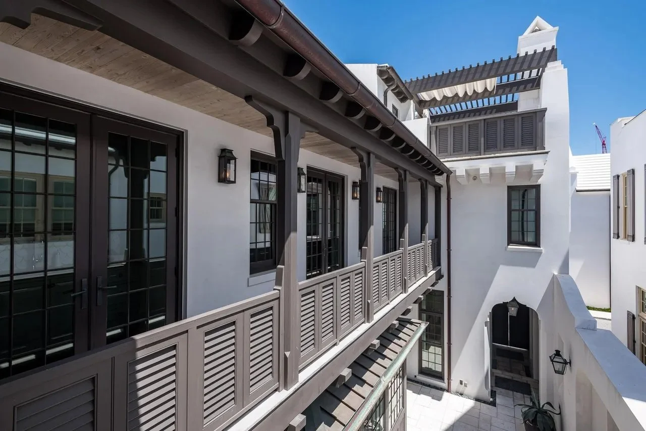View of a white building with multiple windows, balconies with wooden railings, and a small courtyard on a sunny day.