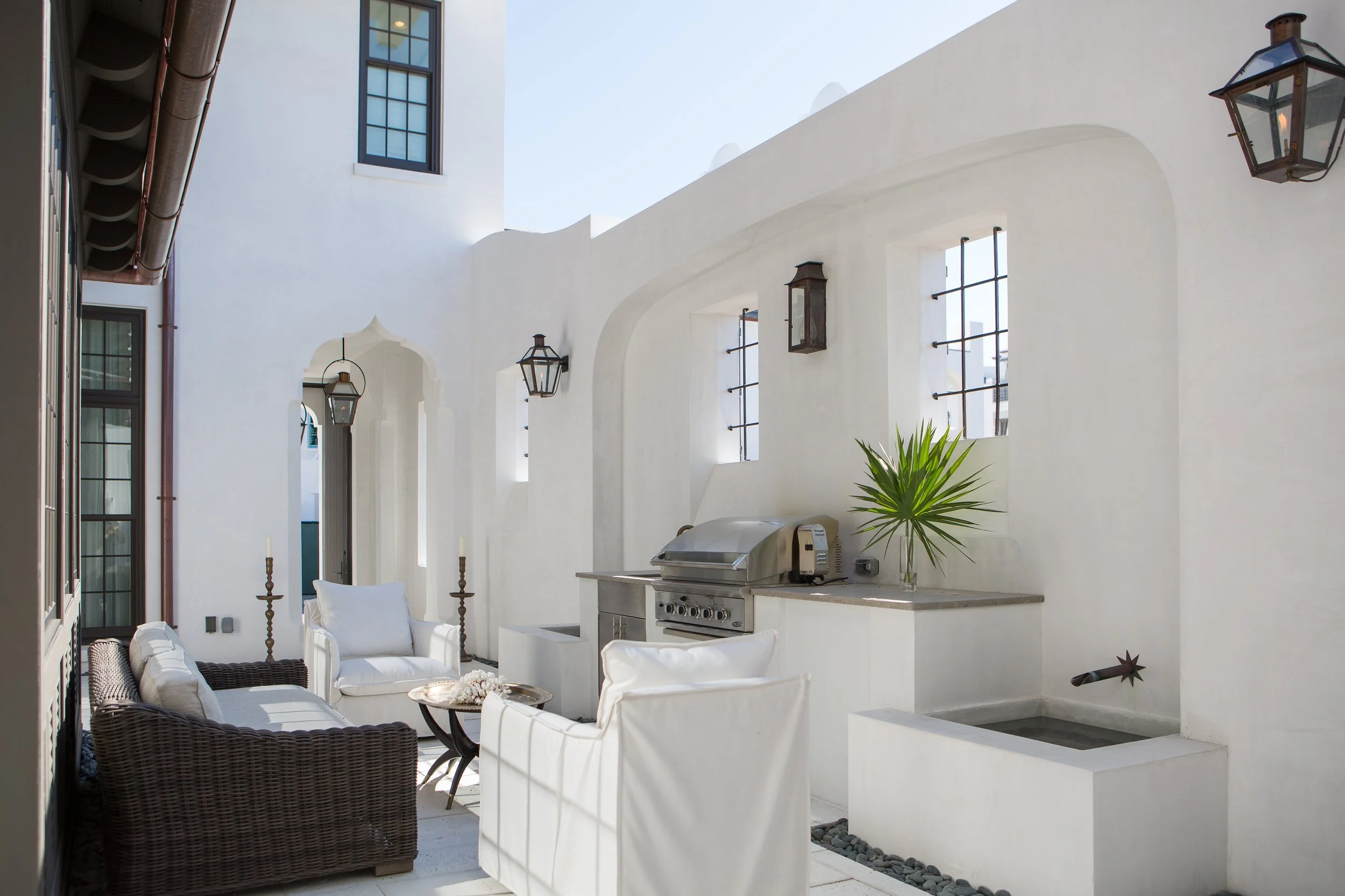 Outdoor patio with white walls, wicker and white cushioned seating, a stainless steel grill, and a potted plant. There are black metal lanterns and a small water feature on the right.