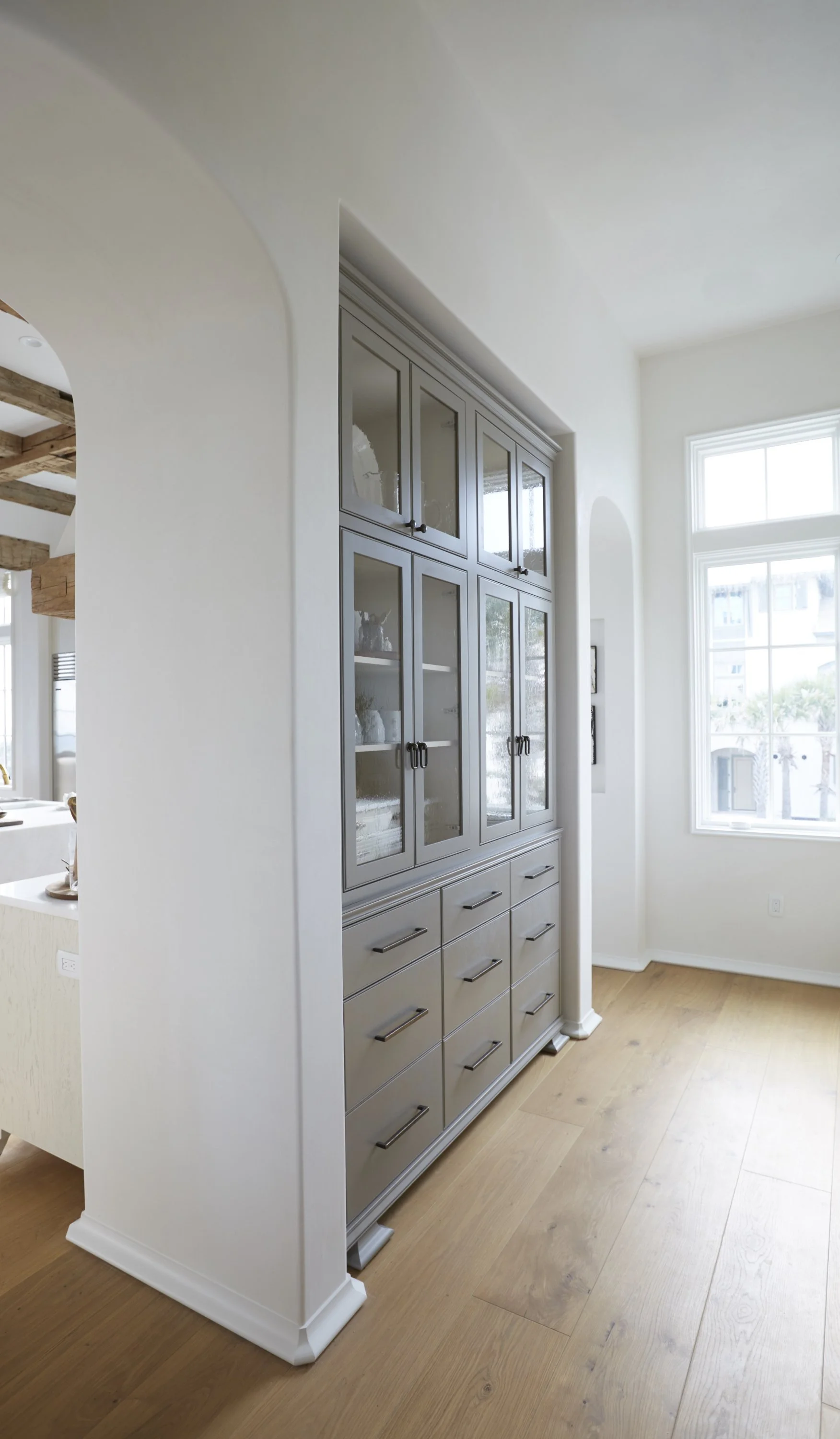 A tall grey display cabinet with glass doors and multiple drawers, situated in a bright room with large windows and light wood flooring.
