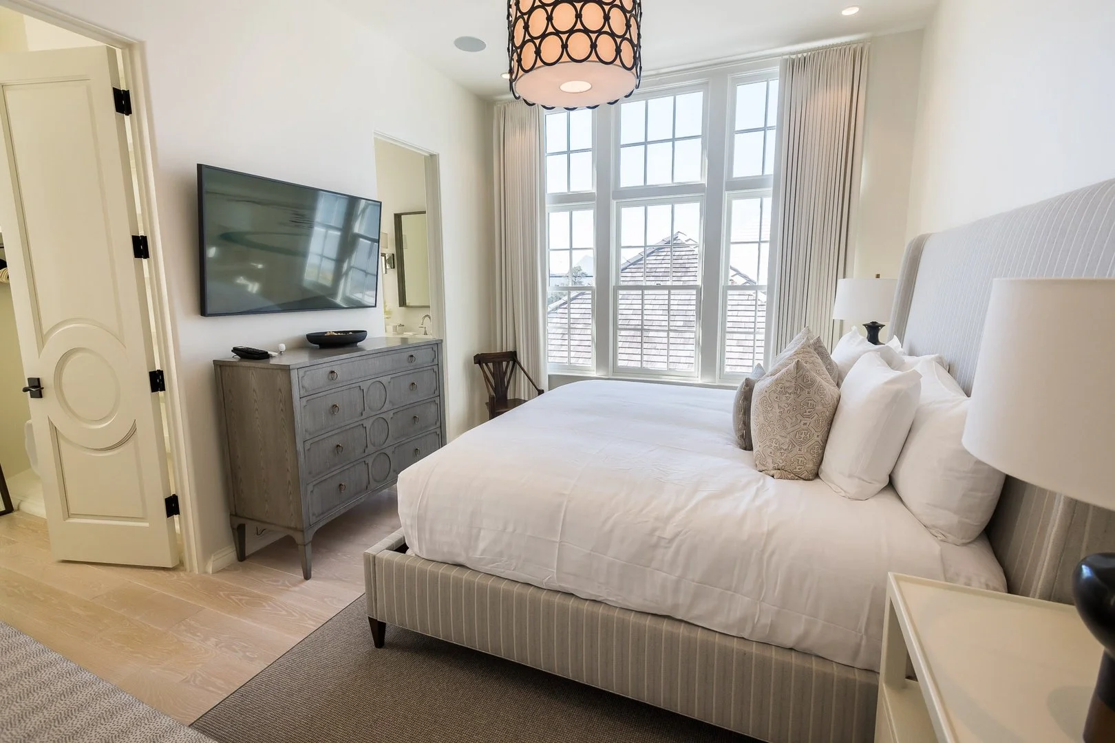A bedroom with a bed, pillows, nightstand, dresser with a TV, and large windows with curtains.