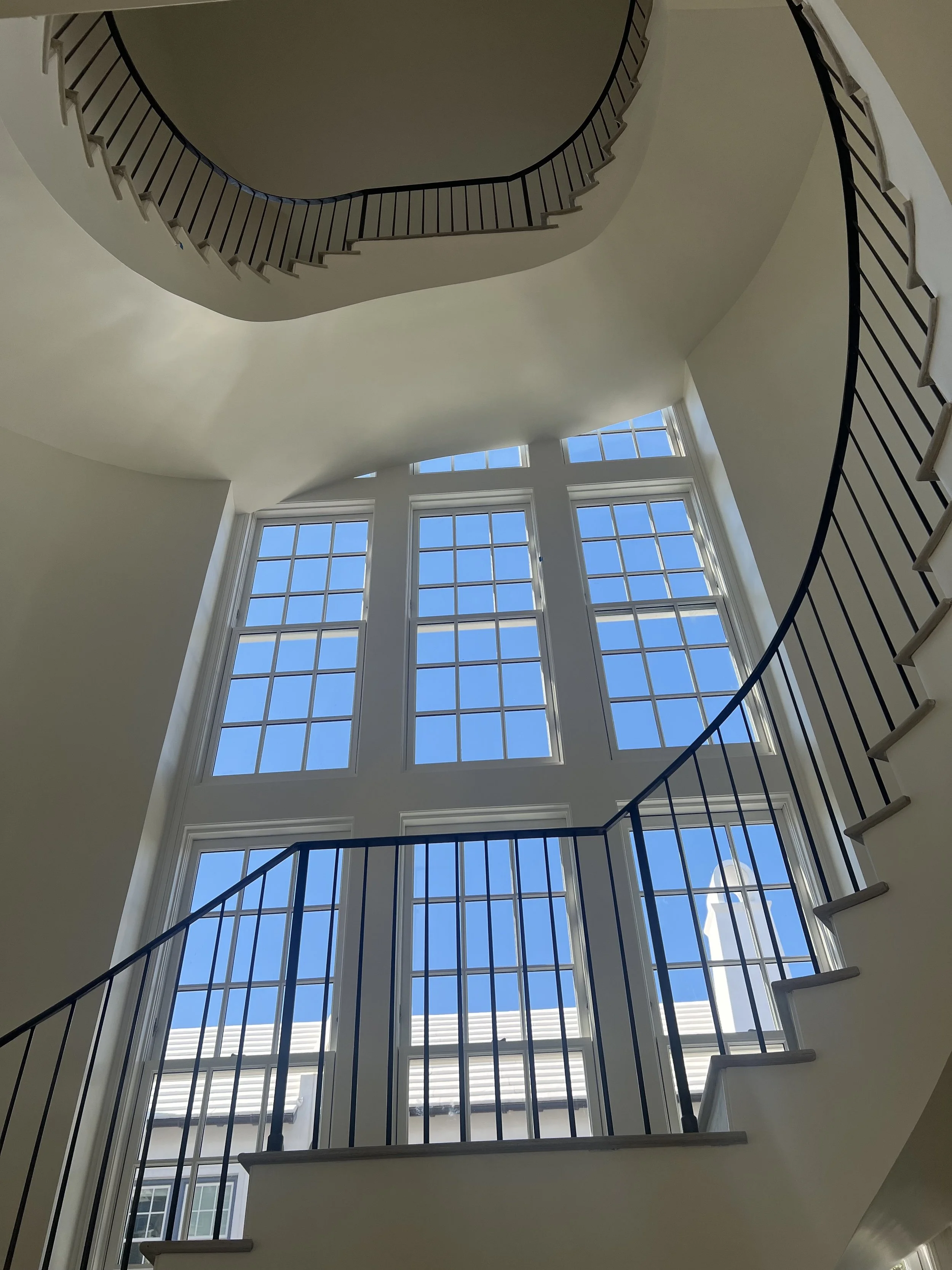 Interior view of a staircase with black metal railings, large multi-pane windows showing a bright blue sky outside, and a curved architectural ceiling.