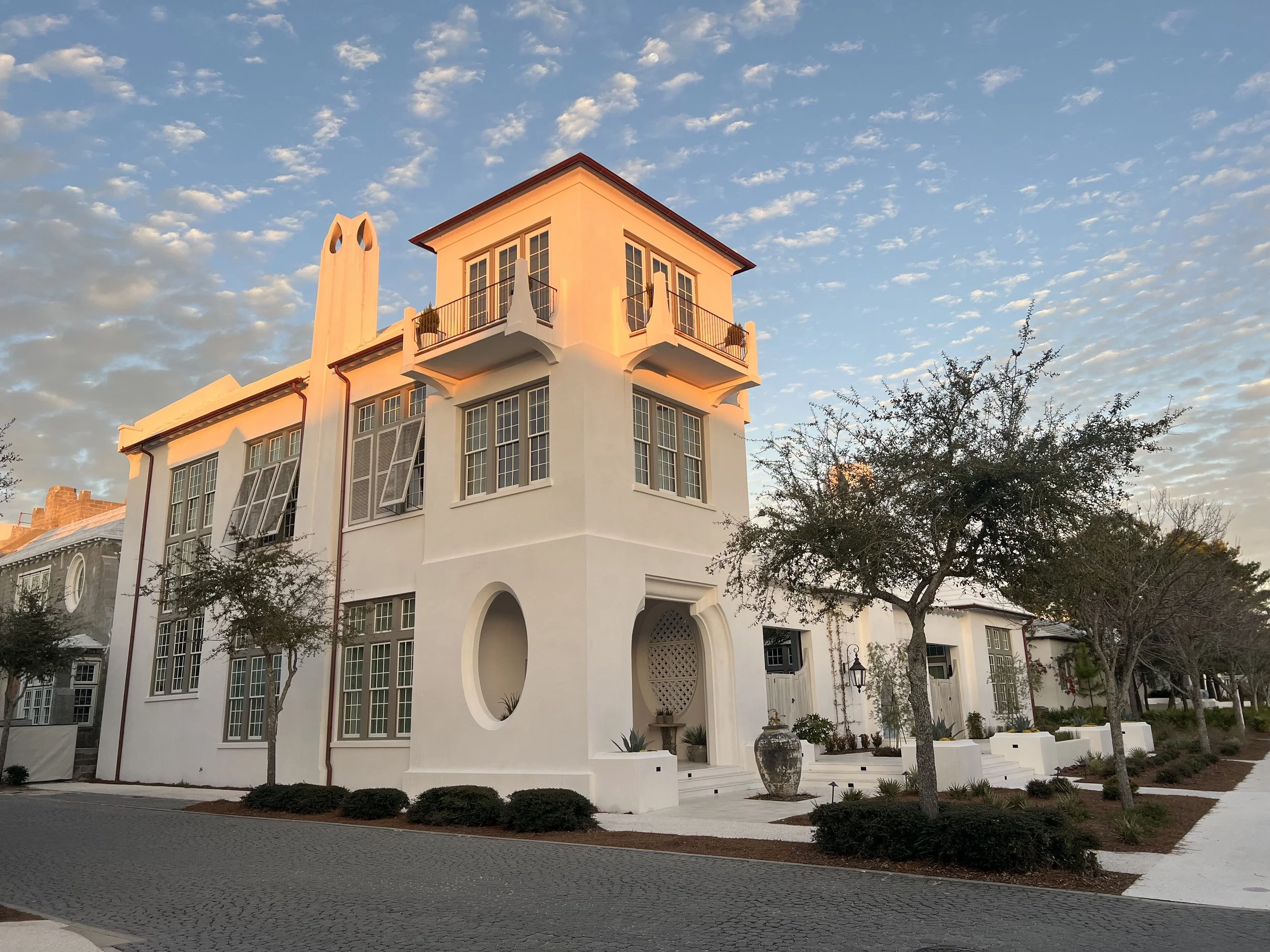 White multi-story house with rounded windows, small balconies, tall chimney, and desert landscaping, under a sky with scattered clouds.