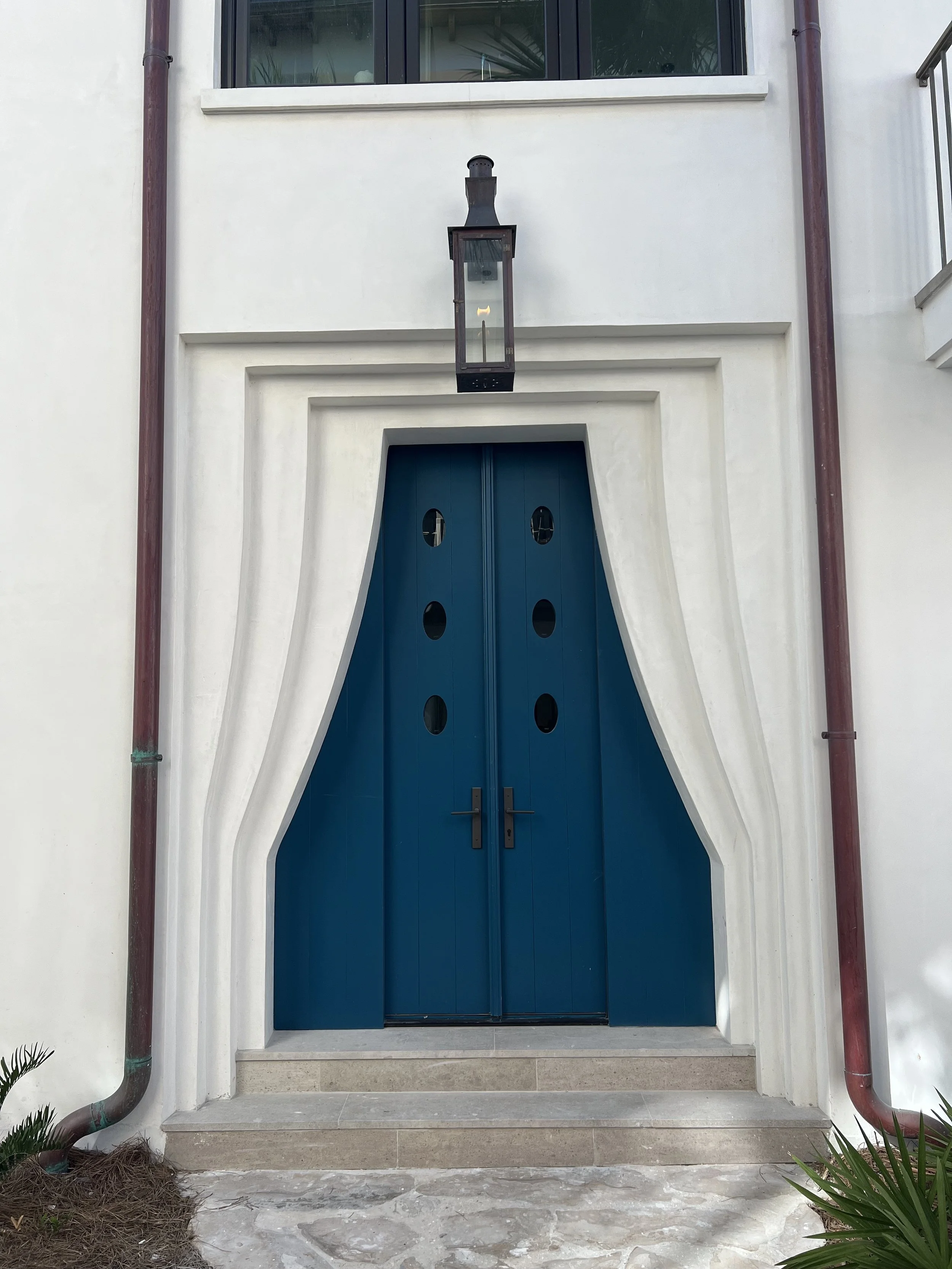 A blue door with a unique, curved white frame on a white building exterior, flanked by brown rain gutter pipes, with a black lantern hanging above the door and a window above that.