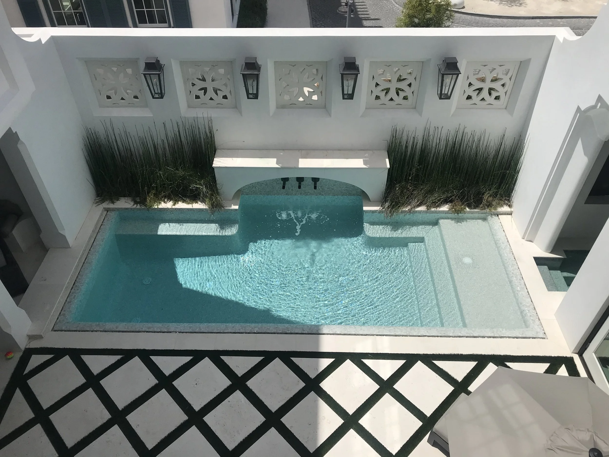 A white outdoor pool area with a small pool, surrounded by white walls with decorative panels and black lantern lights. Tall grass plants line the back wall, and there is a black-and-white patterned floor in the foreground.