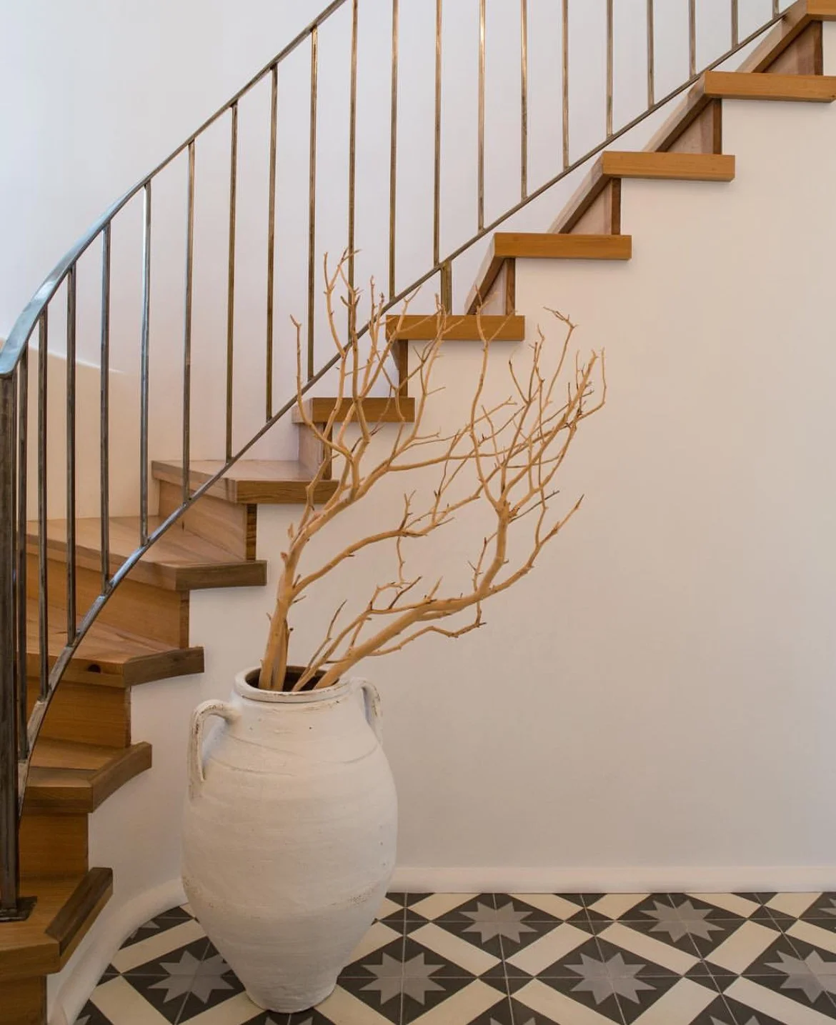 Interior scene with a white wall, a staircase with wooden steps and a metal railing, a floor with black and white patterned tiles, and a large white vase holding a leafless, twisted branch sitting on the floor.