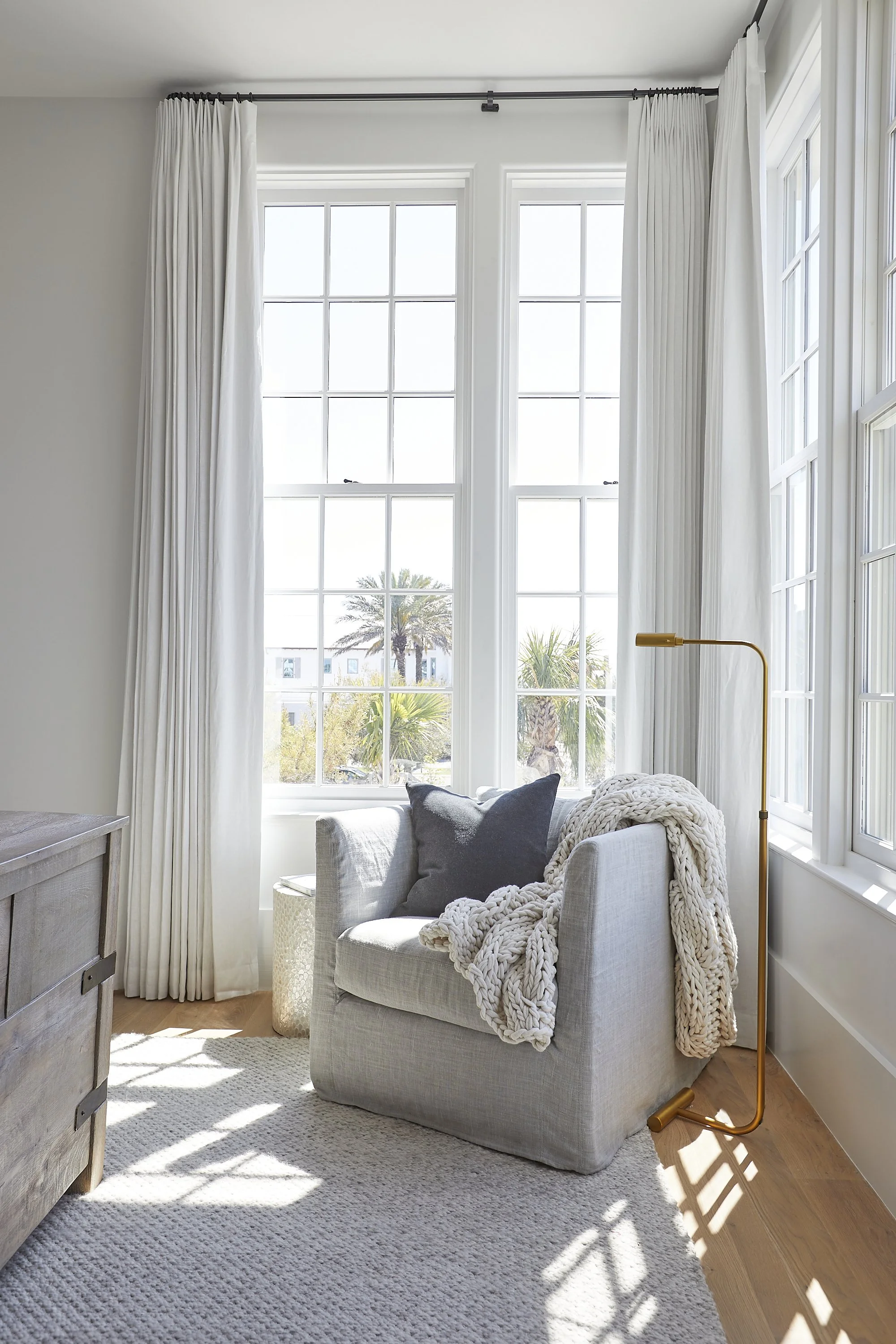 Sunlit living room corner with large white framed windows, a white armchair with dark and light pillows, a chunky knit blanket, a gold floor lamp, a light-colored textured rug, and a wooden side table.
