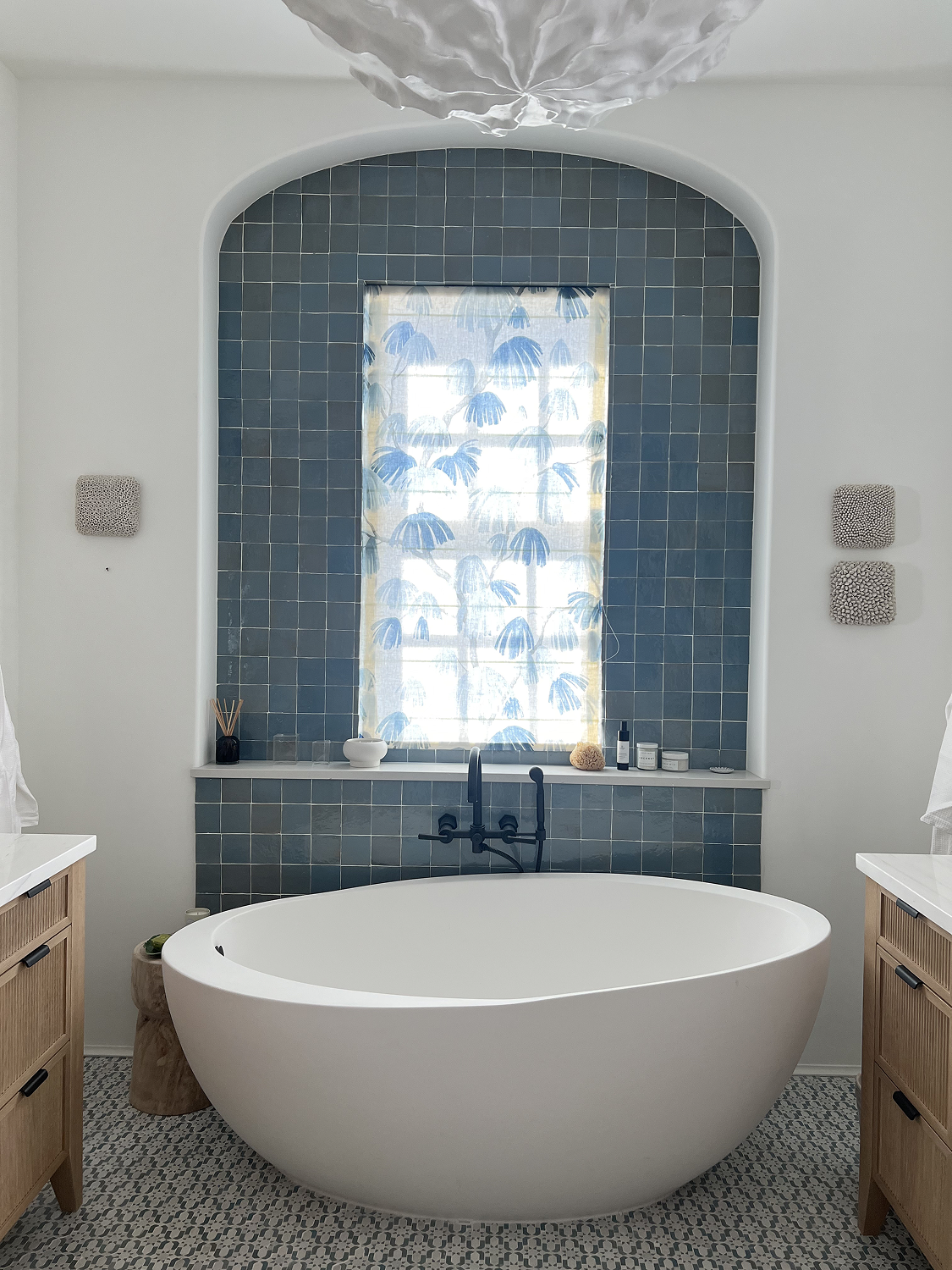 A modern bathroom with a white freestanding bathtub in front of a window with blue palm print curtains. The wall behind the window is tiled in blue, and there are two wooden vanities on either side. The bathroom has a patterned tile floor.