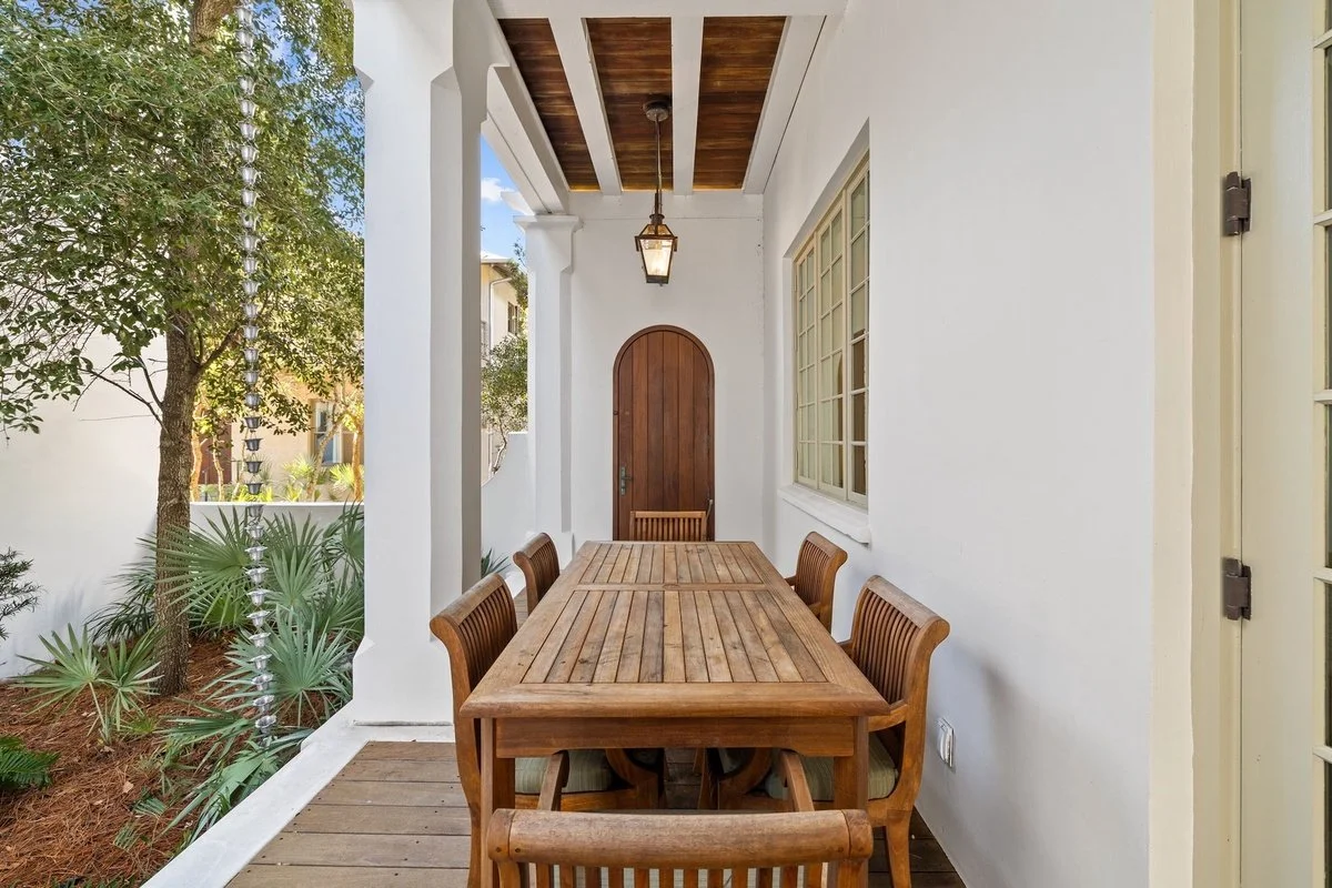 Outdoor porch with a wooden dining table and six matching chairs, white walls, a wooden ceiling with beams, a hanging lantern, and a door at the end of the porch.