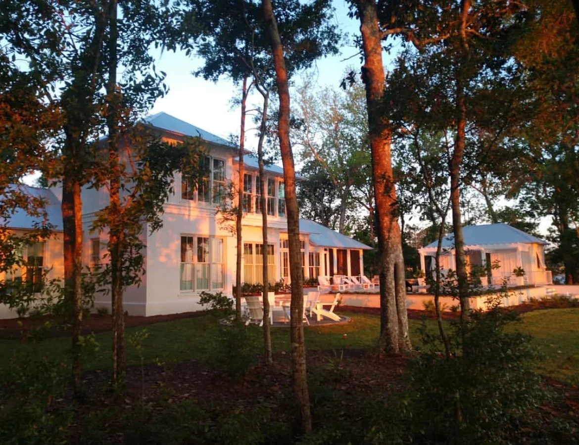 A white house with large windows surrounded by trees, illuminated by warm sunlight, with outdoor patio furniture visible.