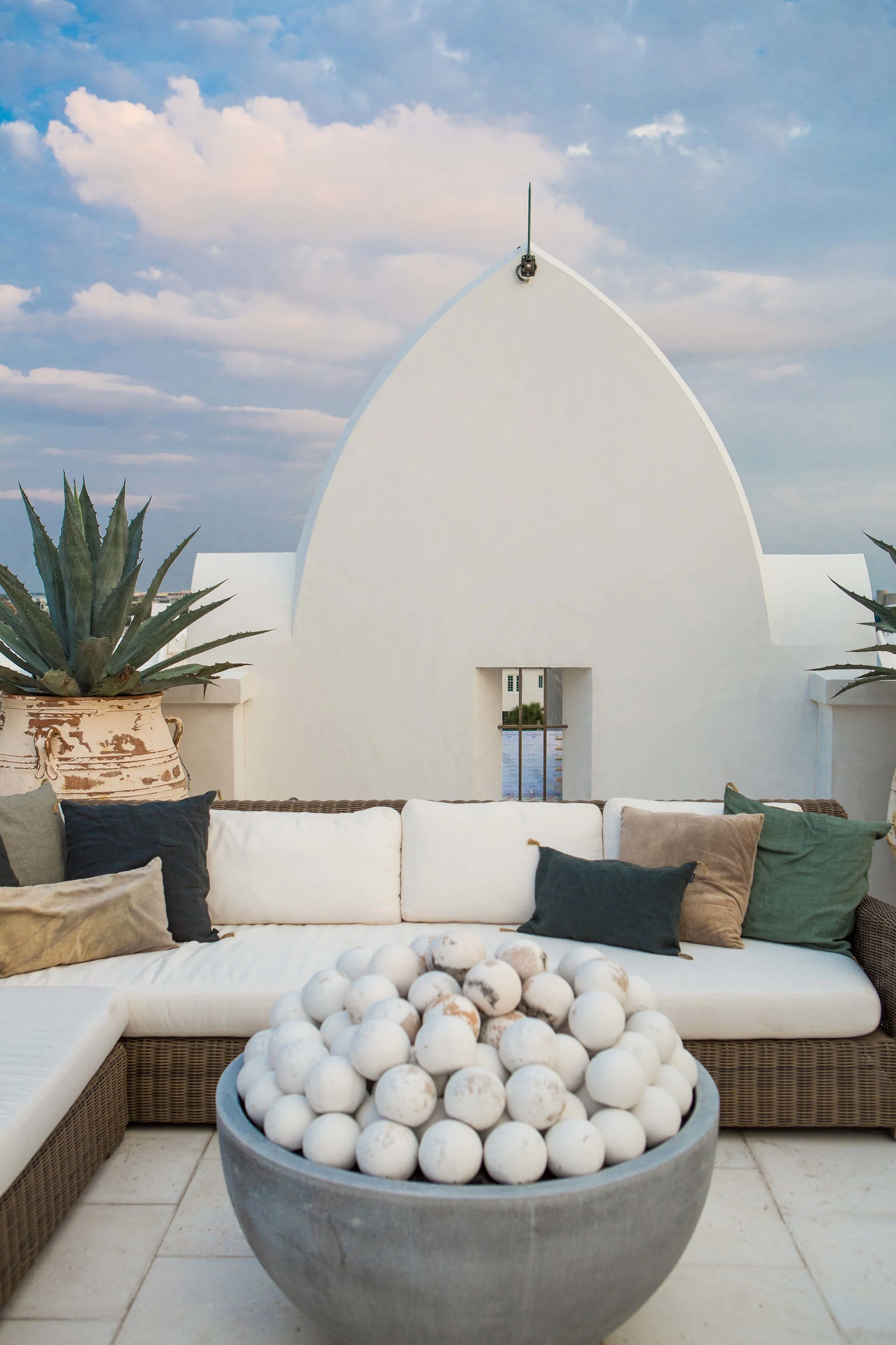 Outdoor seating area with a white cushioned sofa and pillows, a large pot with agave plant, and a bowl of white balls, with a modern white building and a cloudy sky in the background.