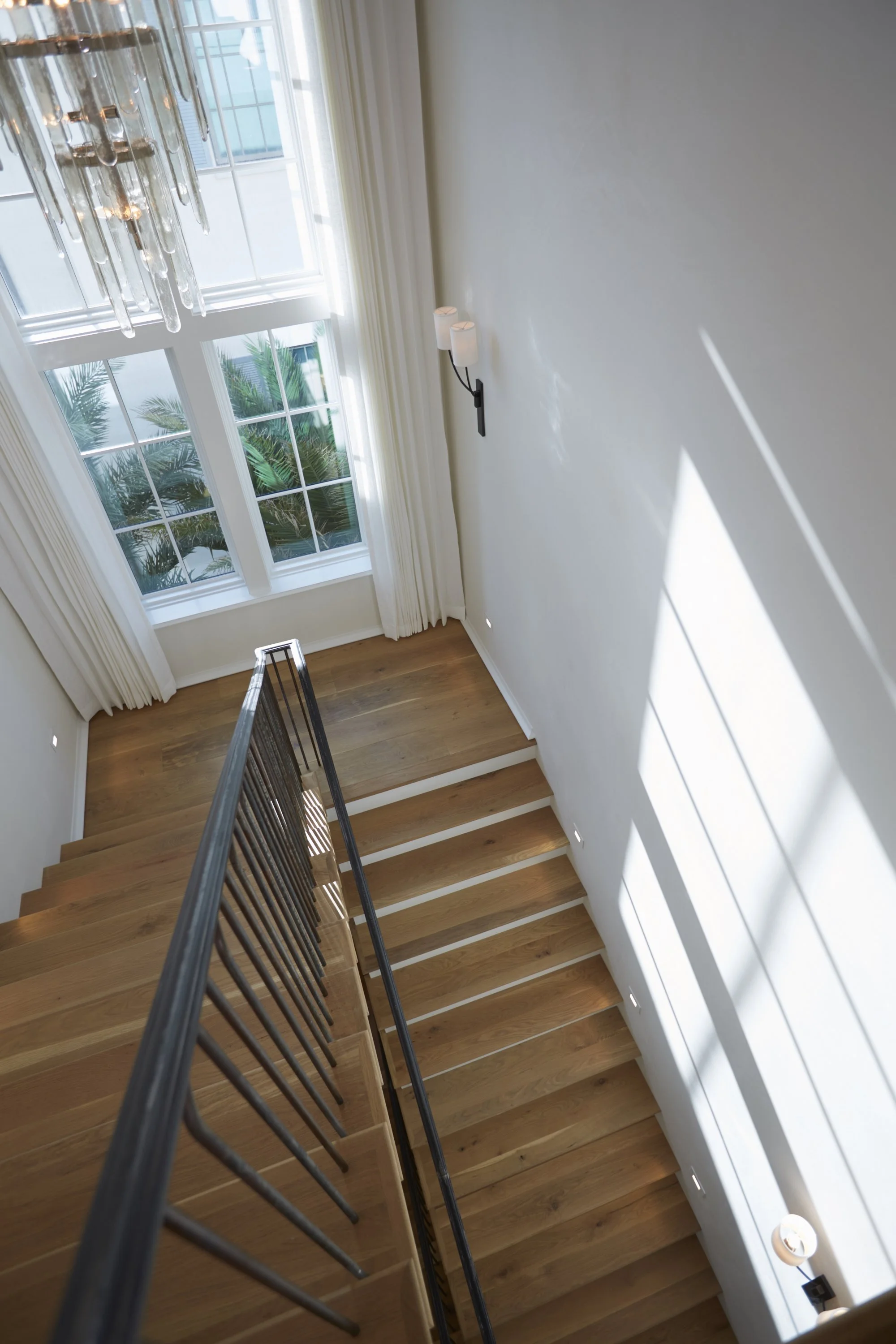 Looking down a staircase with wooden steps, black railing, and a large window at the top, with natural light and greenery outside.