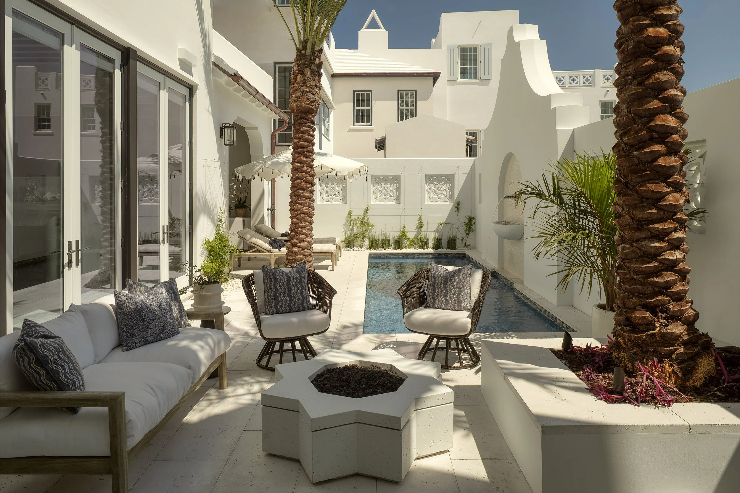 A private outdoor patio with a rectangular pool, white walls, palm trees, lounge chairs with cushions, a sofa with pillows, potted plants, and a fire pit.