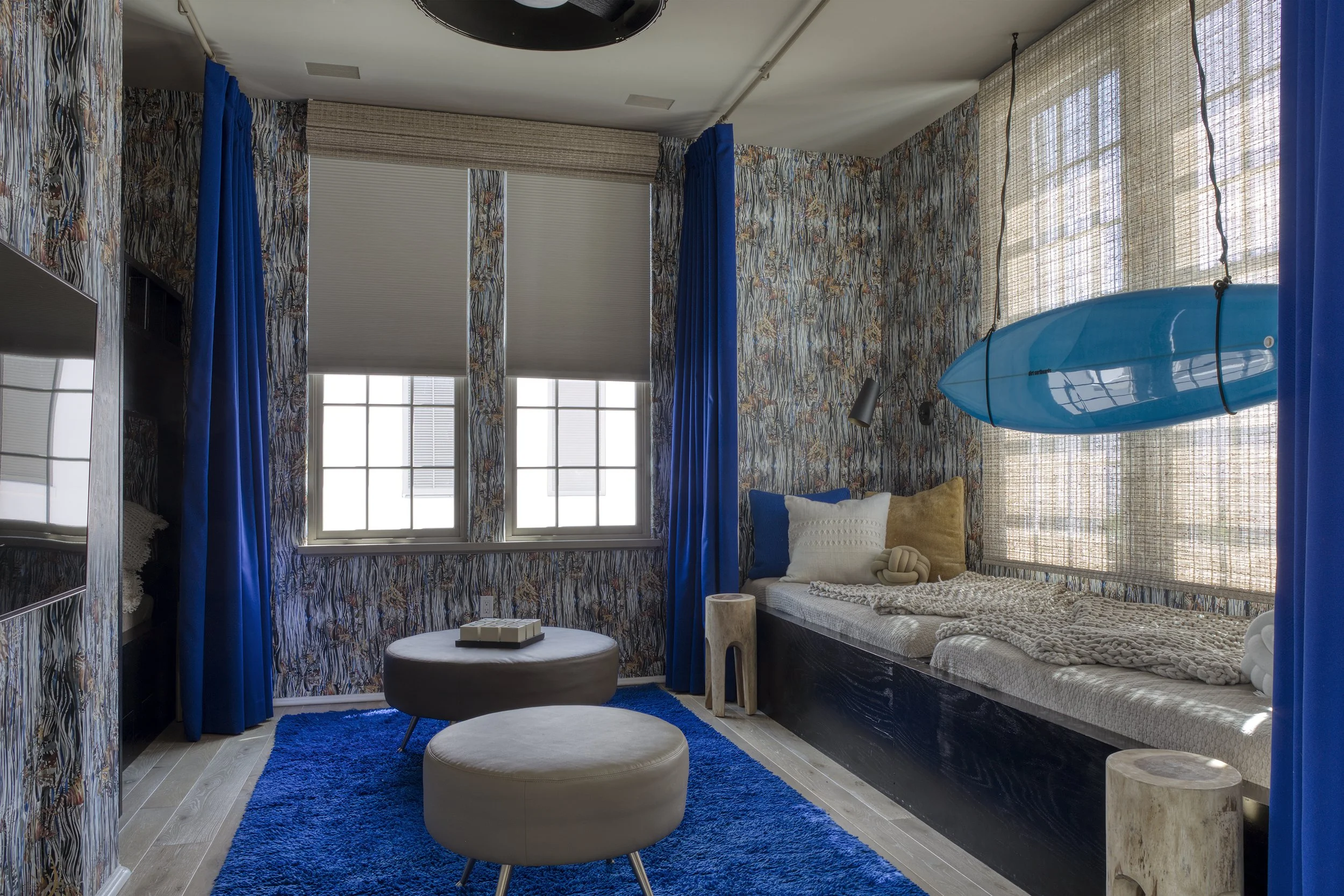 Cozy bedroom with tropical theme, windows with blinds, blue curtains, a blue surfboard hanging from the ceiling, a bed with pillows and knitted throw, two ottomans, a small wooden side table, and a bright blue rug.