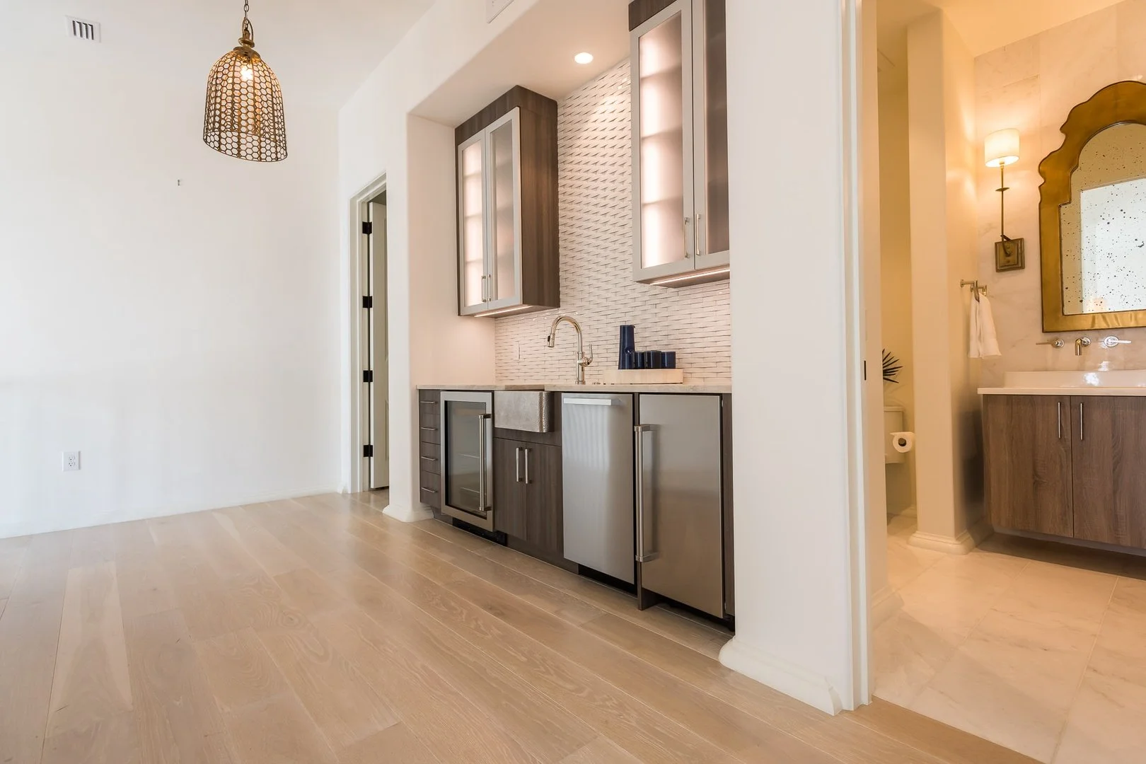 Modern kitchen with gray cabinetry, a small sink, and a textured backsplash, next to a bathroom with a gold-framed mirror, wall-mounted light, and wooden vanity.