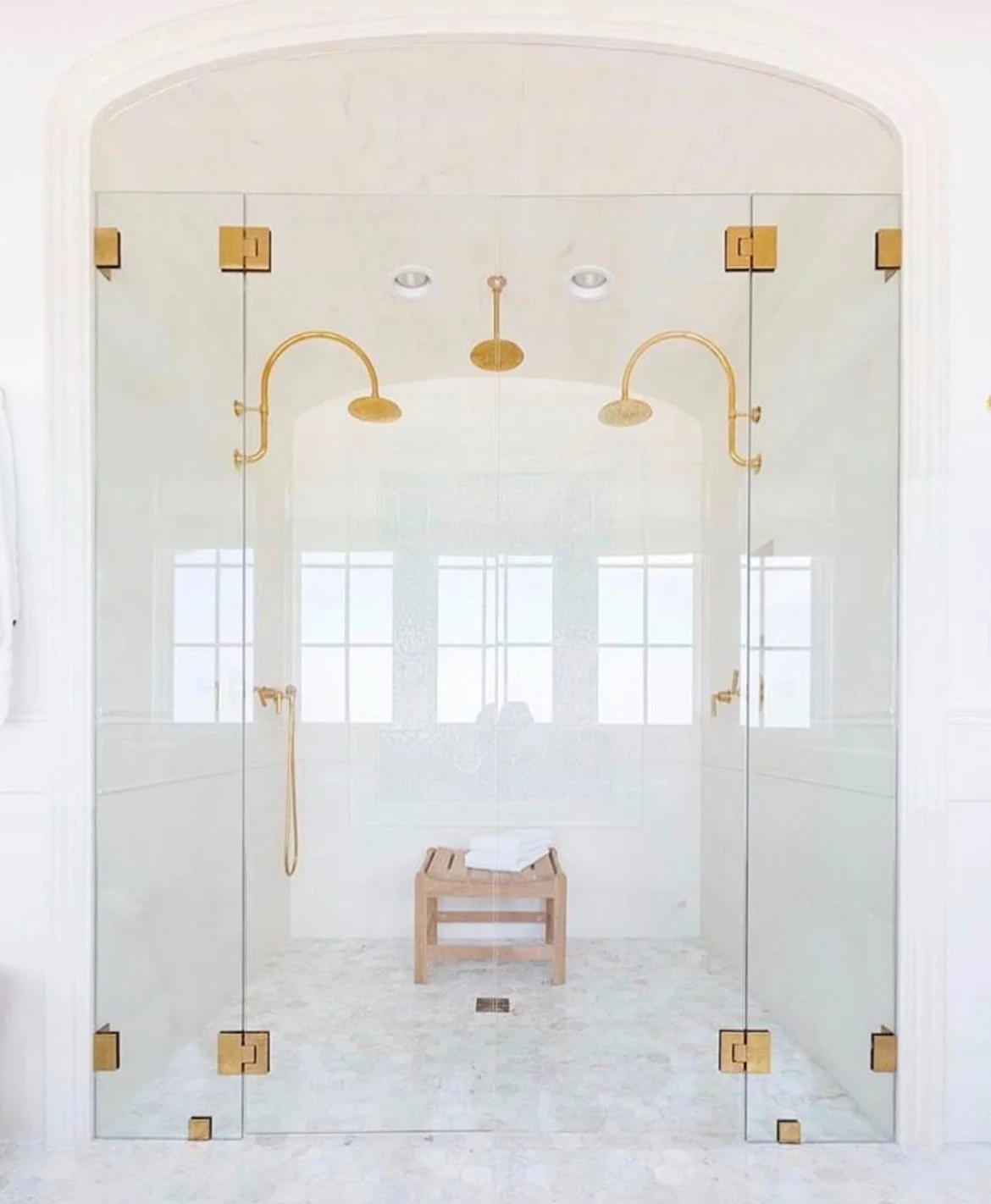 A modern glass shower with gold fixtures, a wooden stool with towels, and natural light from large windows.