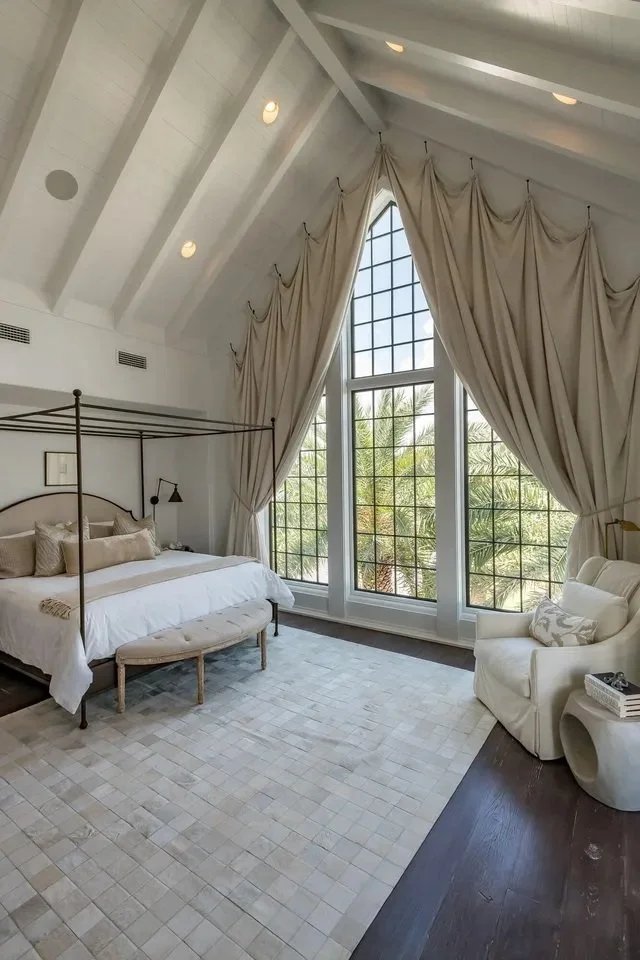 Elegant bedroom with vaulted ceiling, large arched window with cream curtains, white bed with beige pillows, small bench at the foot of the bed, cream armchair, dark wood flooring, and white rug.