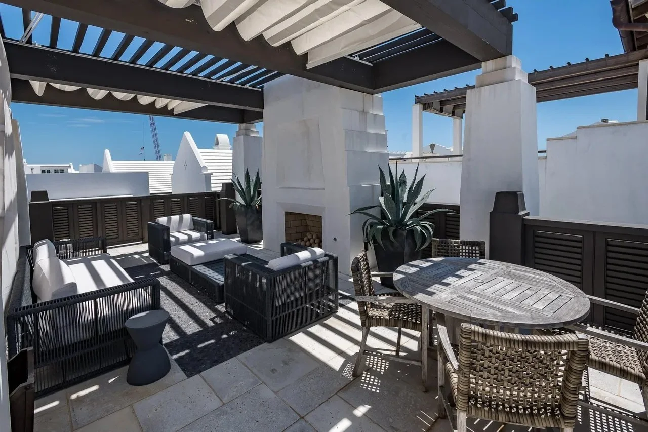 Outdoor rooftop patio with outdoor furniture, large potted plants, a fireplace, and partial shade from a pergola under a clear blue sky.
