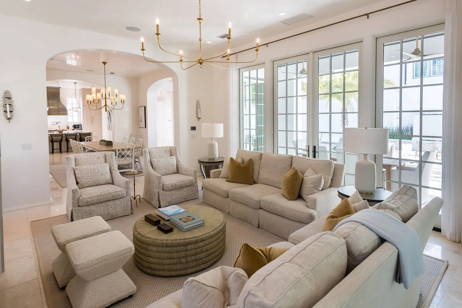Bright living room with cream-colored sofas, patterned armchairs, a textured ottoman, and large glass doors leading outside, decorated with floor lamps and neutral tones.