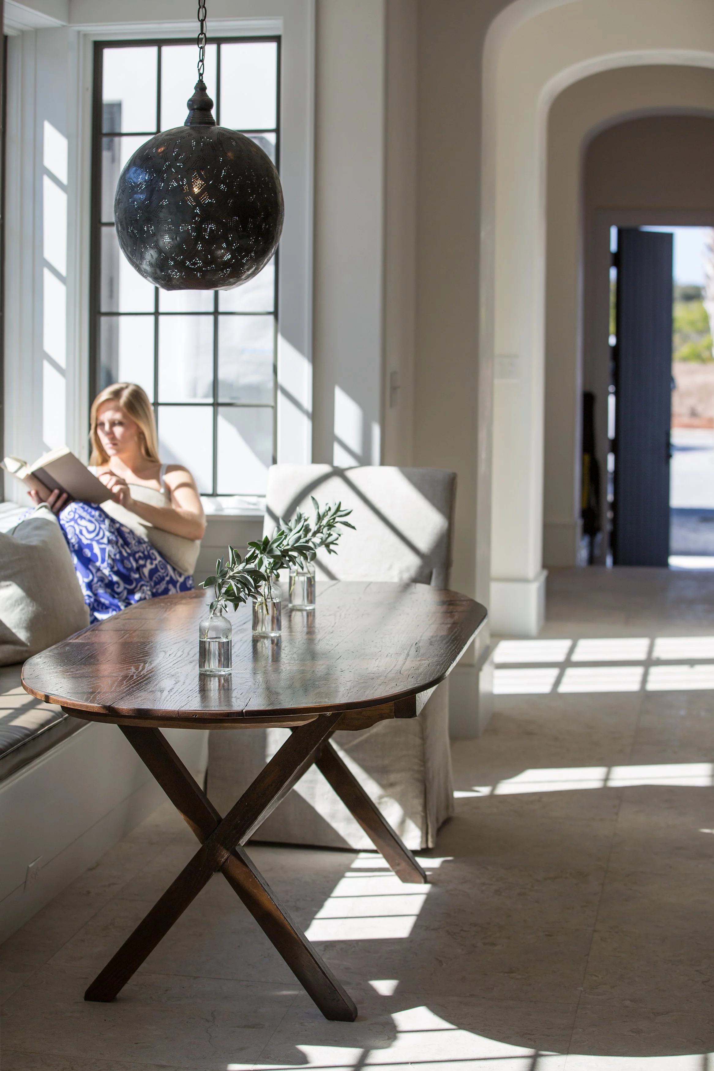 Bright dining area with large window, a hanging black textured pendant light, a wooden table with small glass vases and greenery, and a girl sitting by the window reading a book.