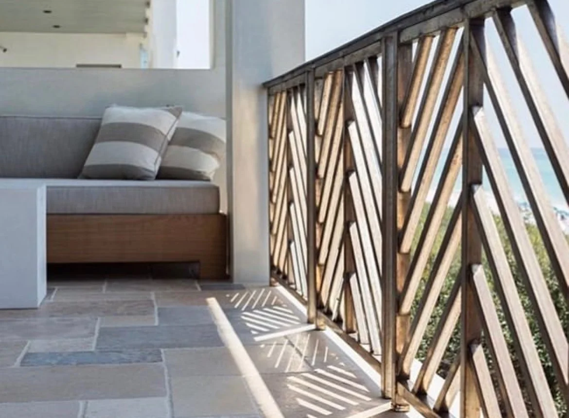 Balcony with a sofa, striped pillows, and a decorative wooden railing with sunlight creating shadows on the tiled floor.