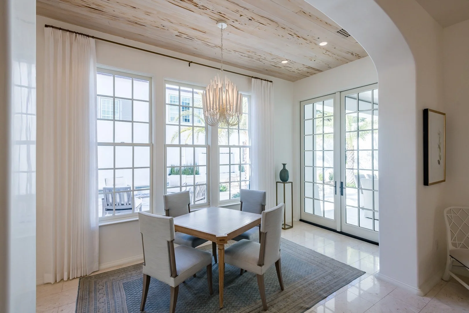 Dining room with a wood dining table and six light-colored upholstered chairs, large windows with white curtains, patio doors, a chandelier, and a decorative vase on a stand.