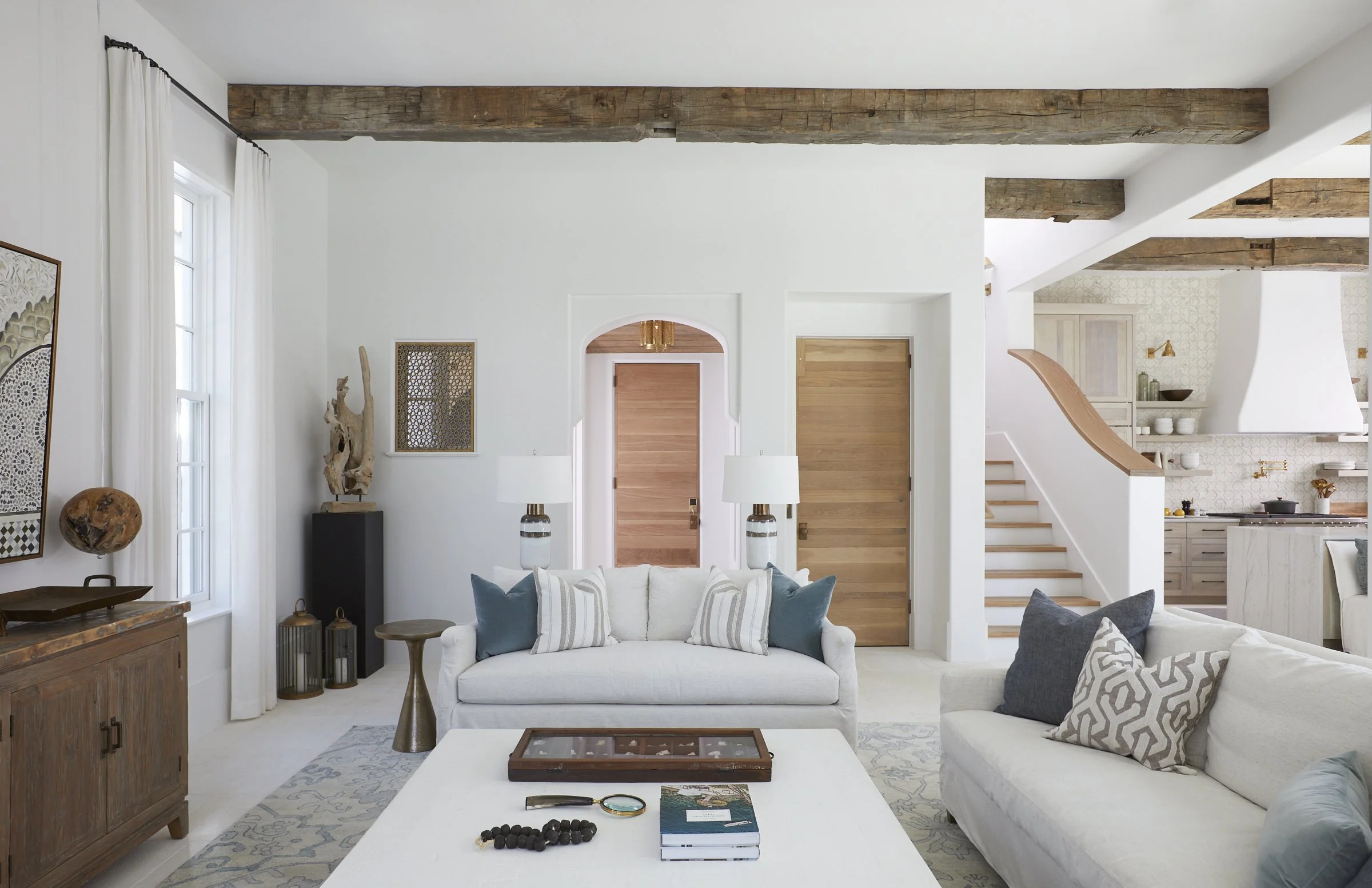 Living room with white couches, wooden beams, and a staircase leading to an upper level, with a kitchen visible in the background.