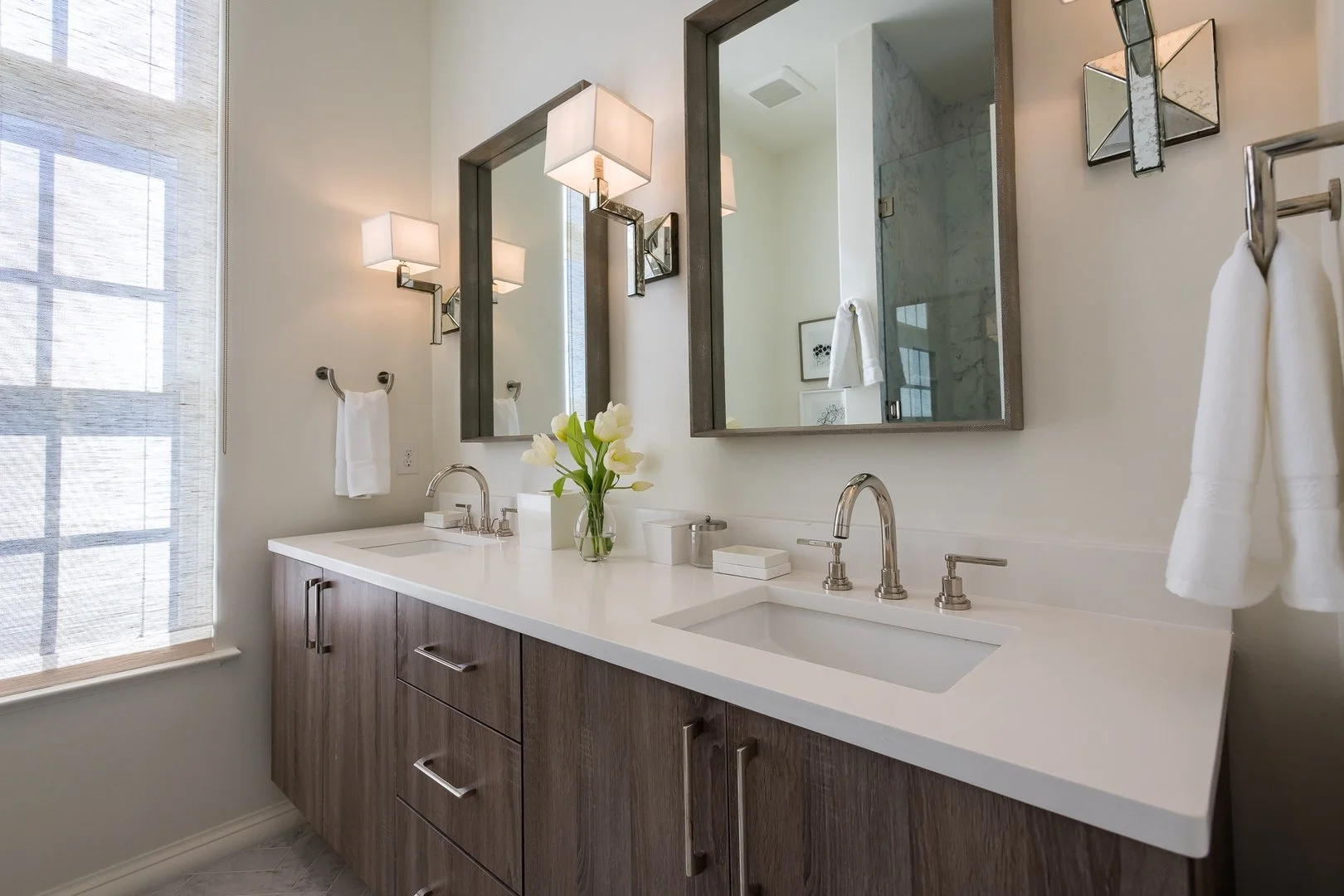 Modern bathroom vanity with dual sinks, large mirrors, wall-mounted lights, a vase with white flowers, and a window with a sheer curtain.