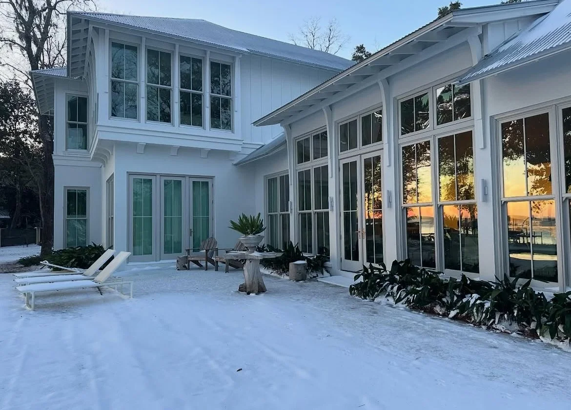 A white house with large windows and a metal roof, surrounded by snow, with a sunset reflecting on the glass, and outdoor lounge chairs and plants on the patio.