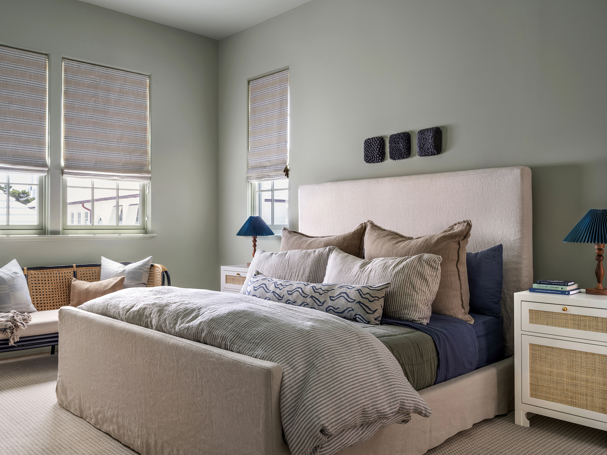 A bedroom with a king-sized bed, multiple pillows, a beige headboard, two white nightstands with blue lamps, and three windows with striped blinds.