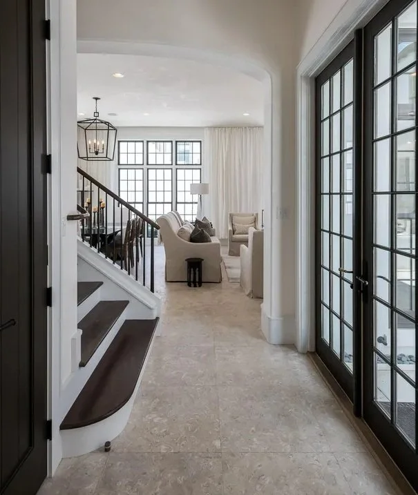 View of a modern living room with large windows, white walls, and beige tile flooring, seen through an entryway with glass doors.