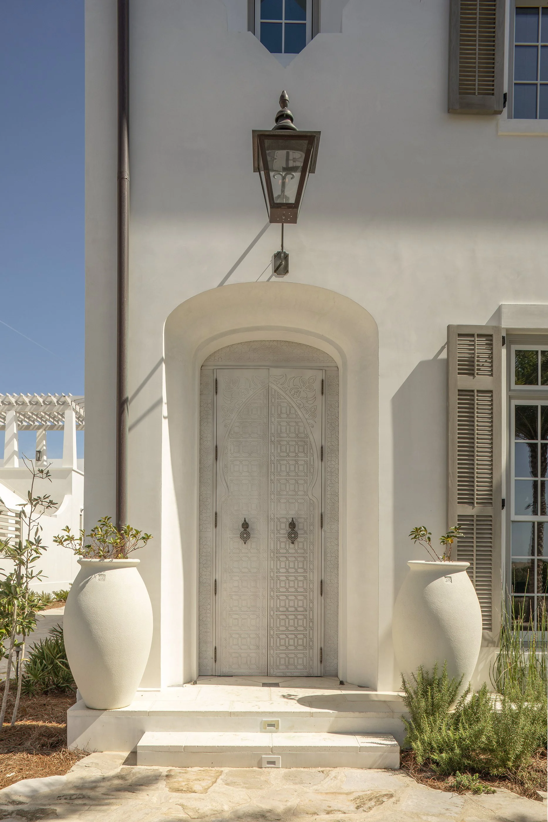 White front door with intricate geometric design, arched surround, flanked by large white pots with plants, and above is an outdoor lantern on a cream-colored wall.