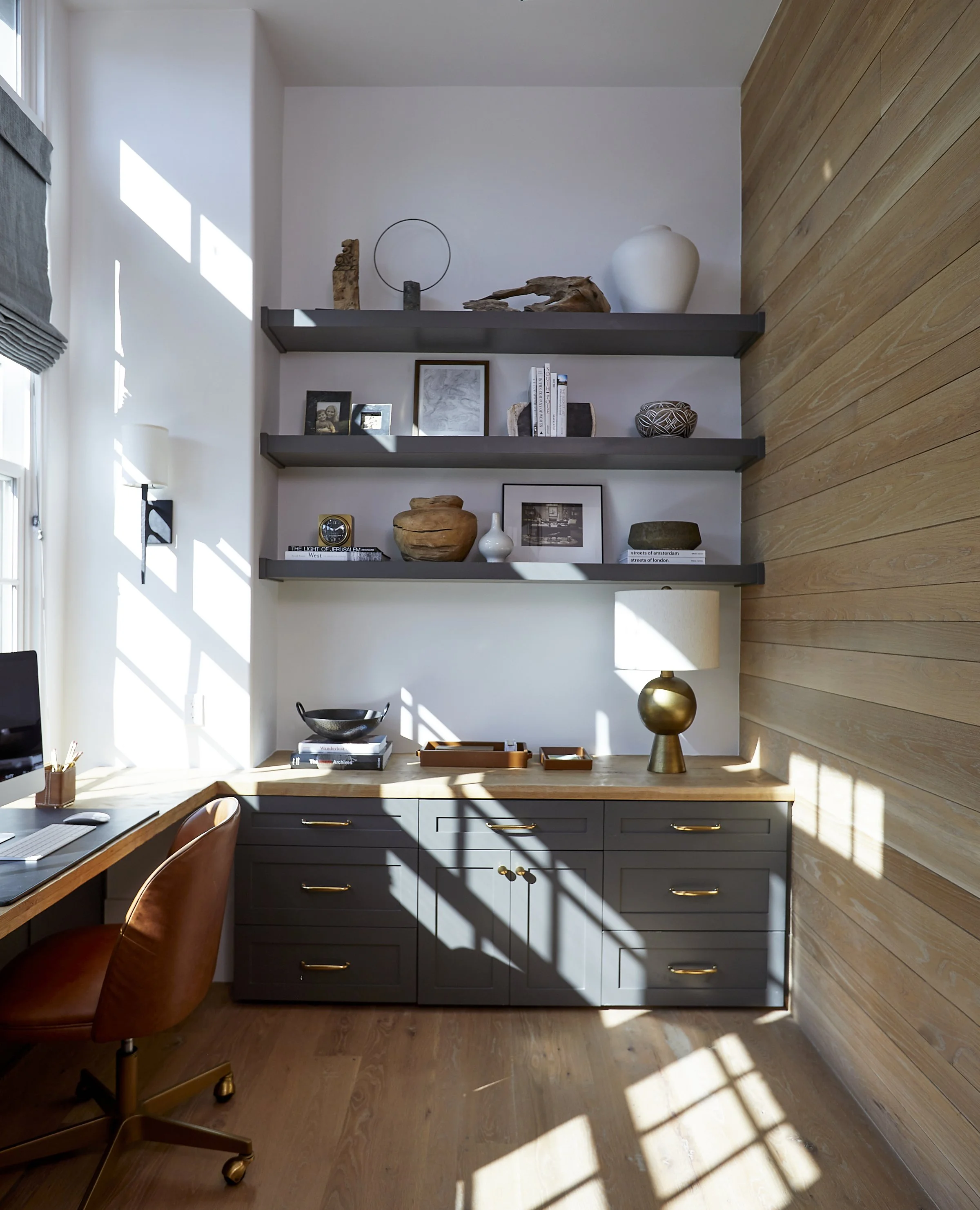 Sunlit home office with wood-paneled wall, grey cabinet, and black floating shelves displaying decorative items and framed photos.