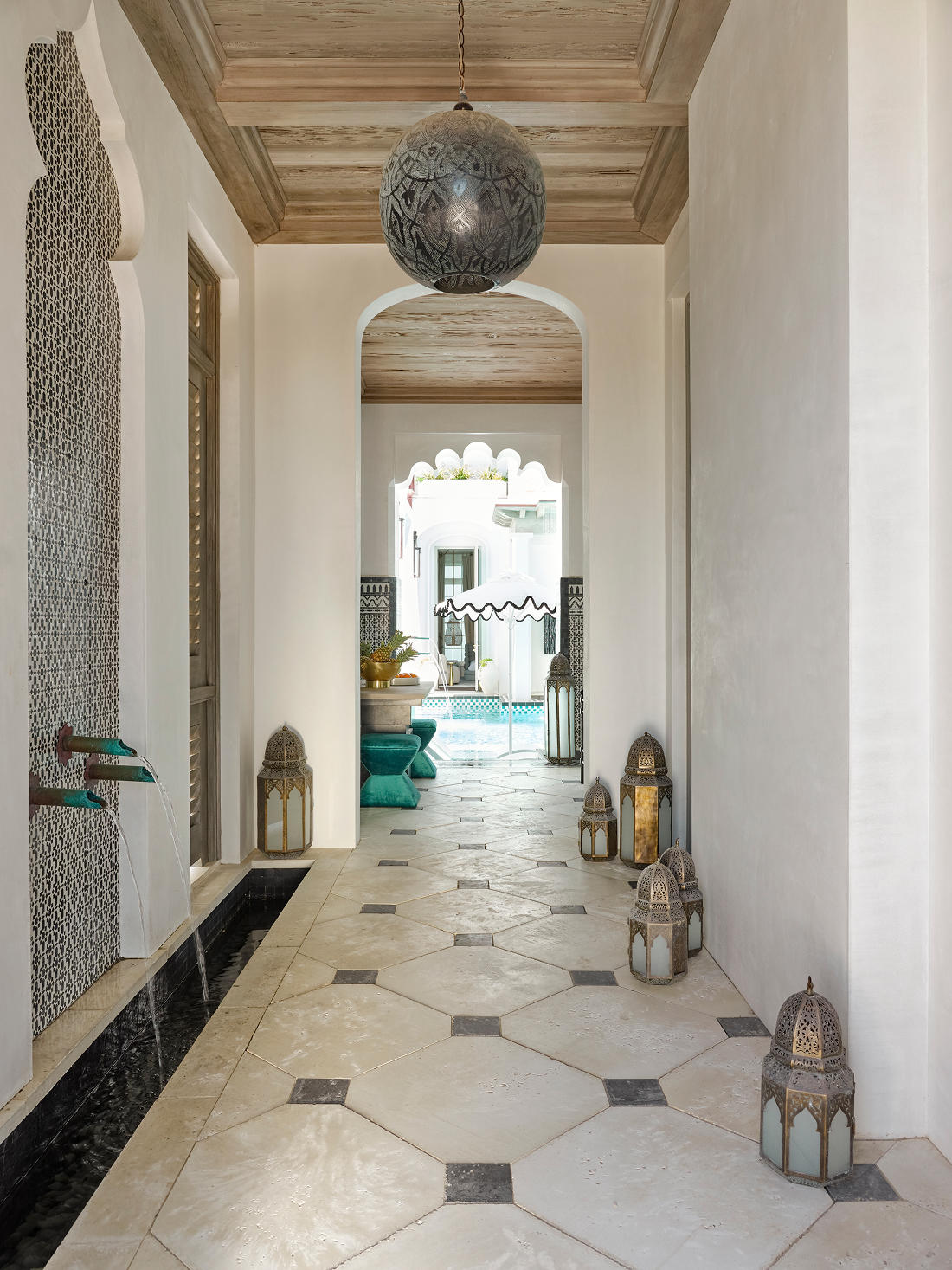 Indoor hallway with a high wooden ceiling, white walls, decorative lanterns along the floor, a large hanging pendant light, and a view of a courtyard with a swimming pool and outdoor furniture in the background.