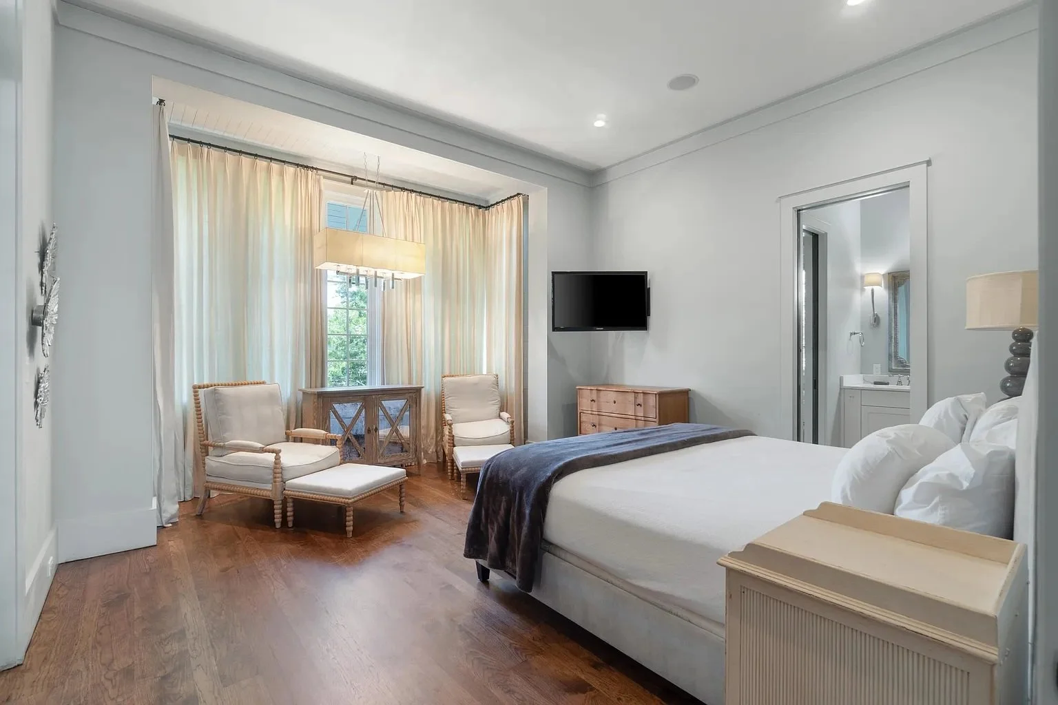 A bedroom with a large bed, white bedding, and pillows, a nightstand, a wall-mounted TV, two chairs near a window, and a bedside lamp, with a bathroom visible in the background.