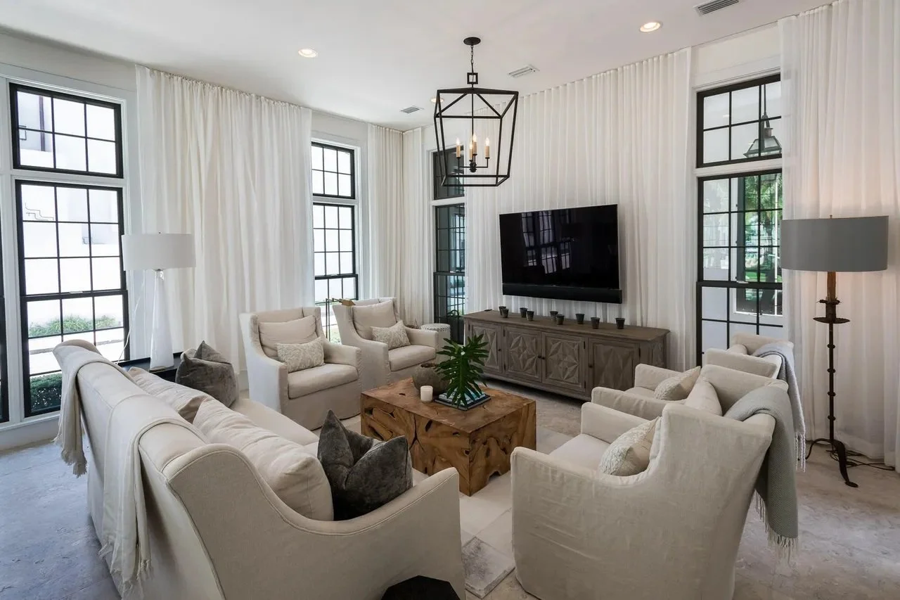 Living room with white curtains, beige sofas and armchairs, a wooden coffee table, a TV mounted on the wall, and large windows with black frames.