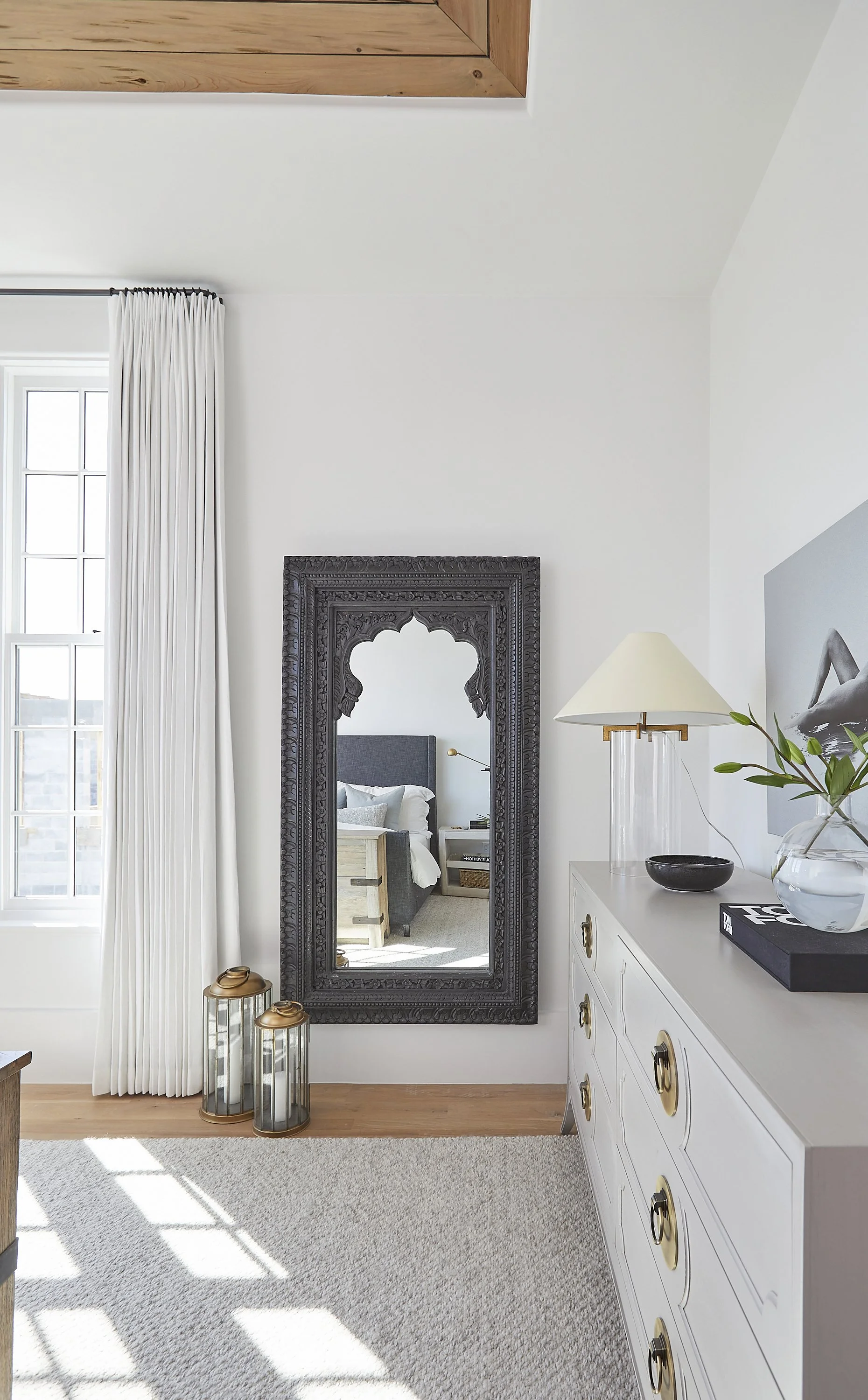 A white dresser with gold handles topped with a lamp, a bowl, and a plant in a vase. A large black decorative mirror and two lanterns placed on the floor next to a window with white curtains. The room has light-colored carpet and wooden flooring.