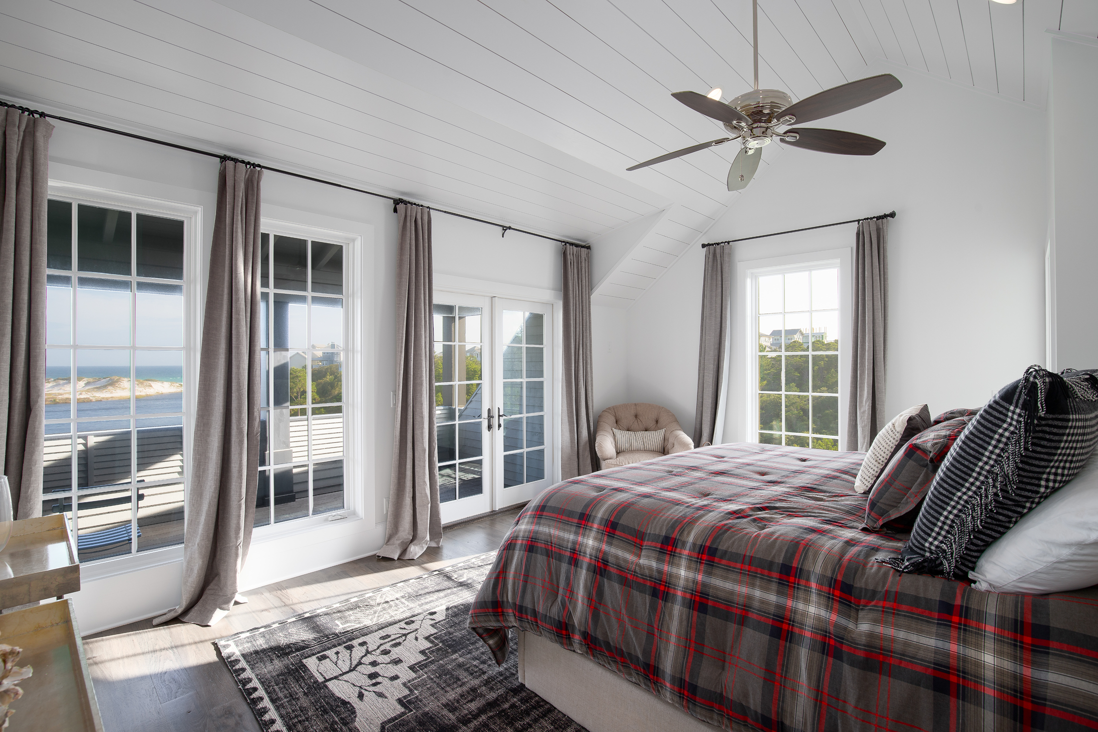 A bright bedroom with large windows showing a beach and ocean view, a bed with plaid bedding, a beige armchair, a ceiling fan, and light-colored curtains.