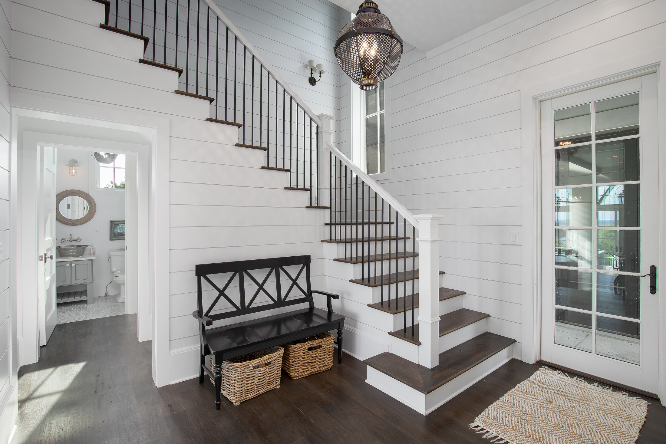 Entryway with wooden stairs and black railing, white shiplap walls, a black bench with baskets underneath, a glass door, a small rug, and a view into a bathroom.