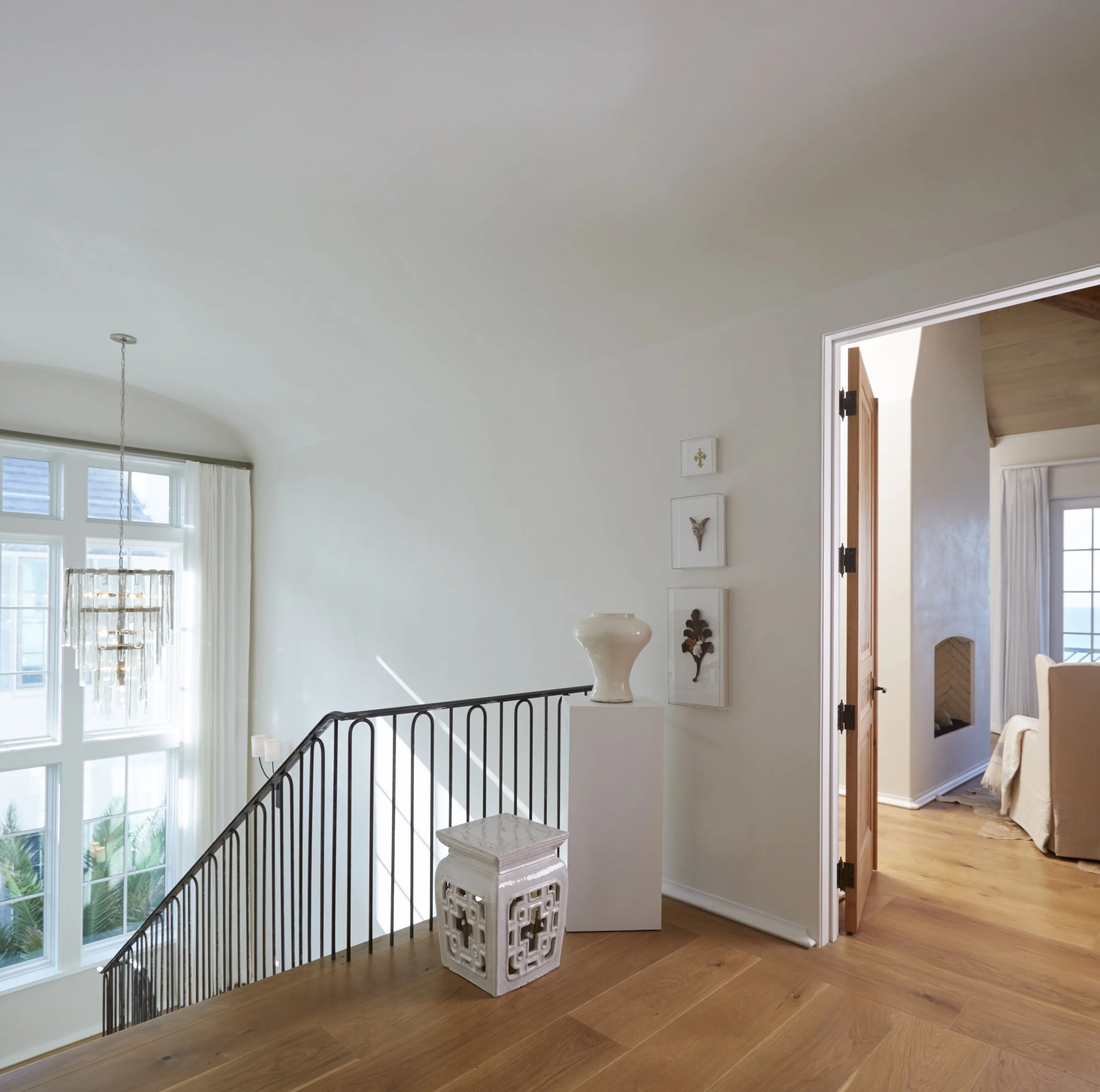 Bright interior of a home with natural wooden floors, white walls, a staircase with black railings, and a hallway leading to a bedroom with beige furniture and large windows.