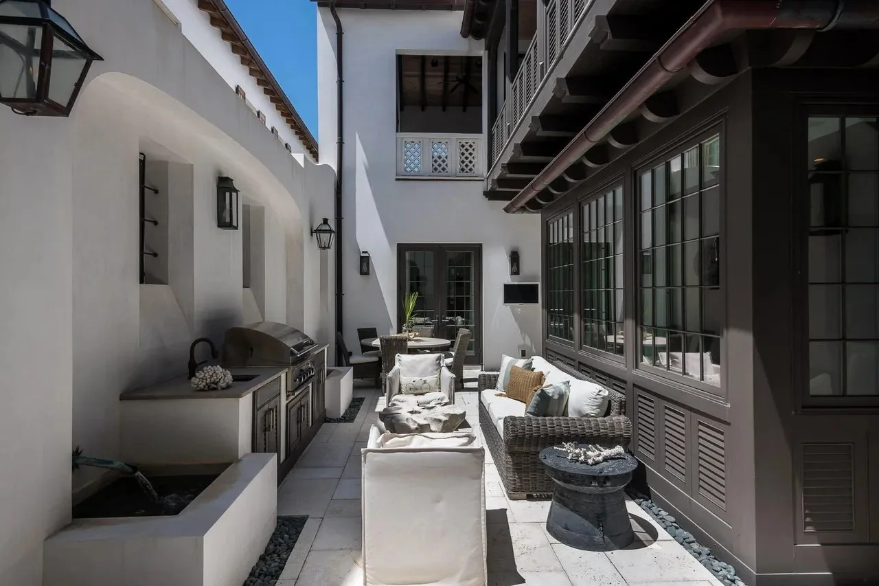 Outdoor patio area with white walls, wicker and fabric seating, a round stone table, potted plants, and a built-in grill under a bright blue sky.