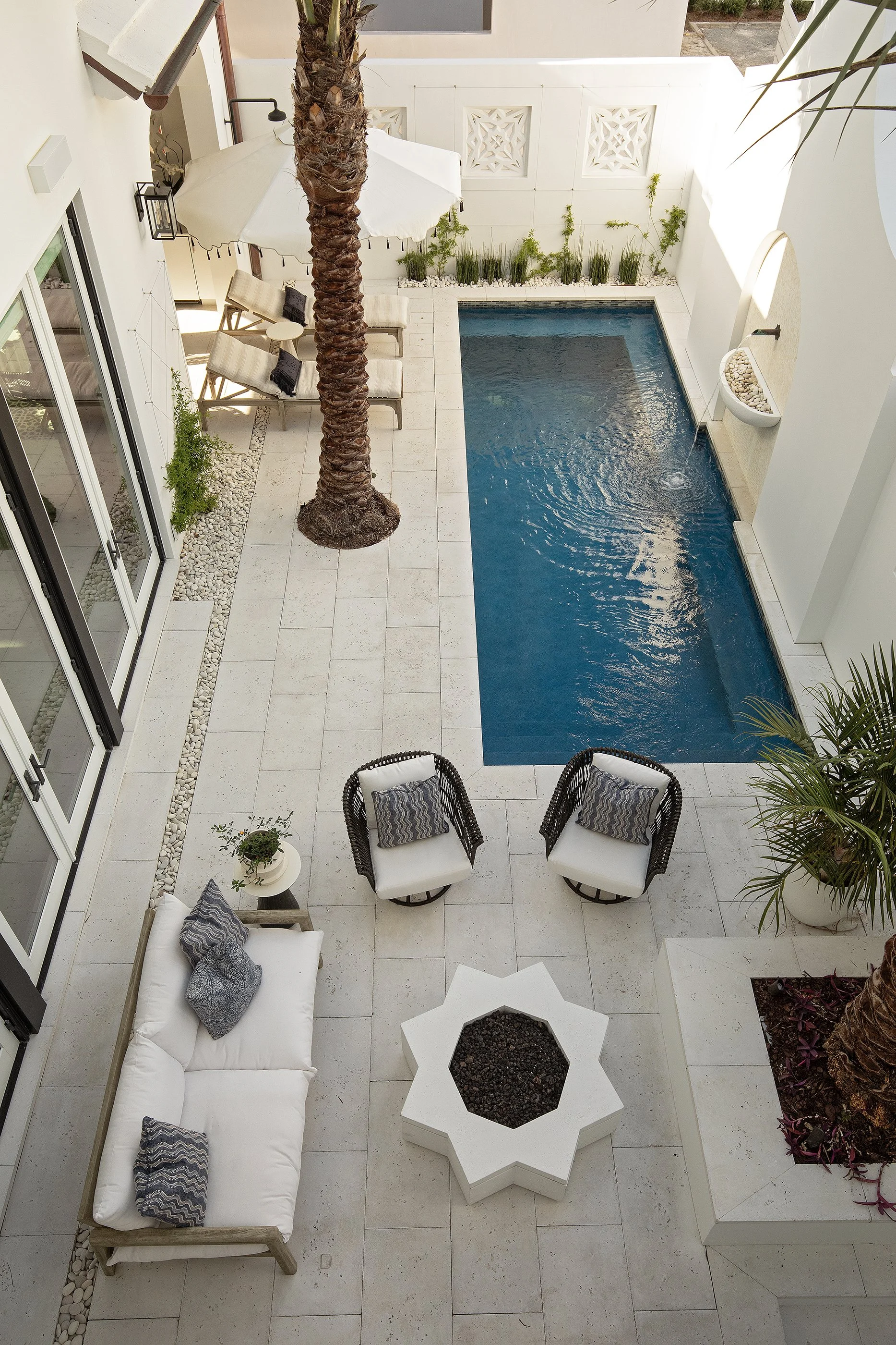 A rooftop patio with a rectangular pool, a white umbrella and lounge chairs, a seating area with a white sofa, striped chairs, a white star-shaped fire pit, and potted plants. The space has white walls and large windows.