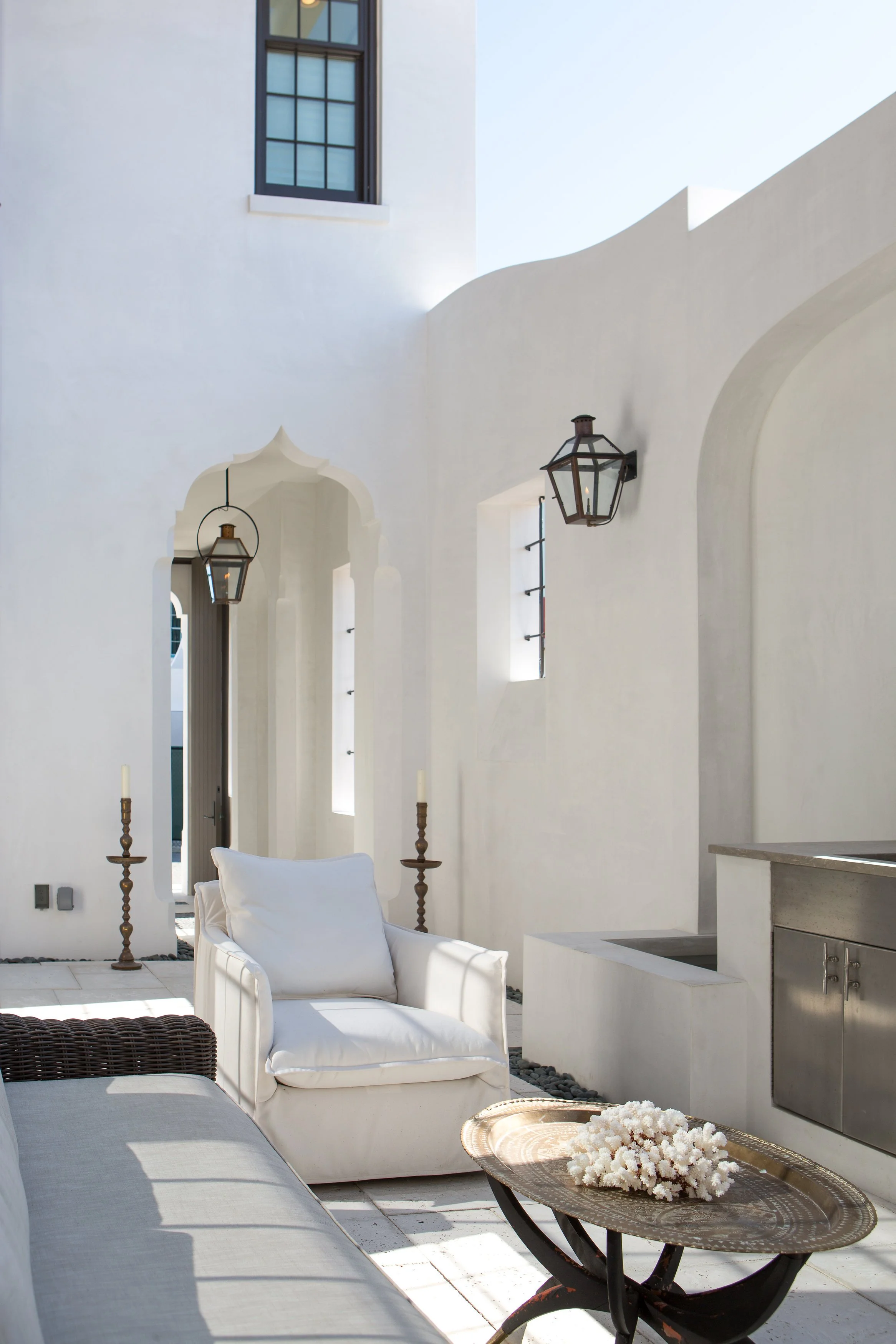 A bright outdoor patio with white walls, a white armchair with a pillow, a wicker sofa, a small table with white flowers, and black lantern-style wall lights.