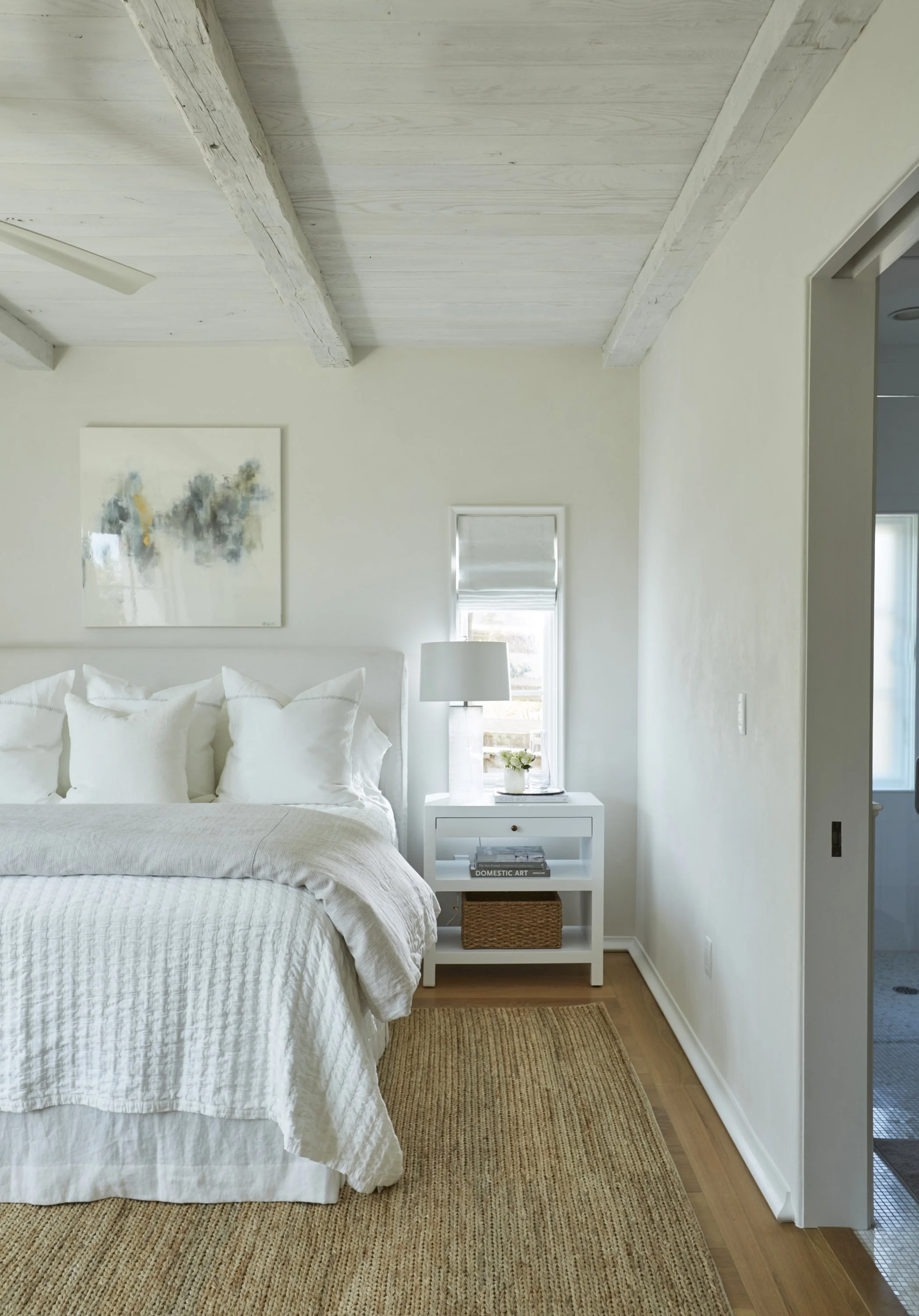 A cozy bedroom with a white bed, multiple pillows, a white bedside table with a lamp, a plant, and books, and artwork on a white wall with exposed wooden beams on the ceiling.