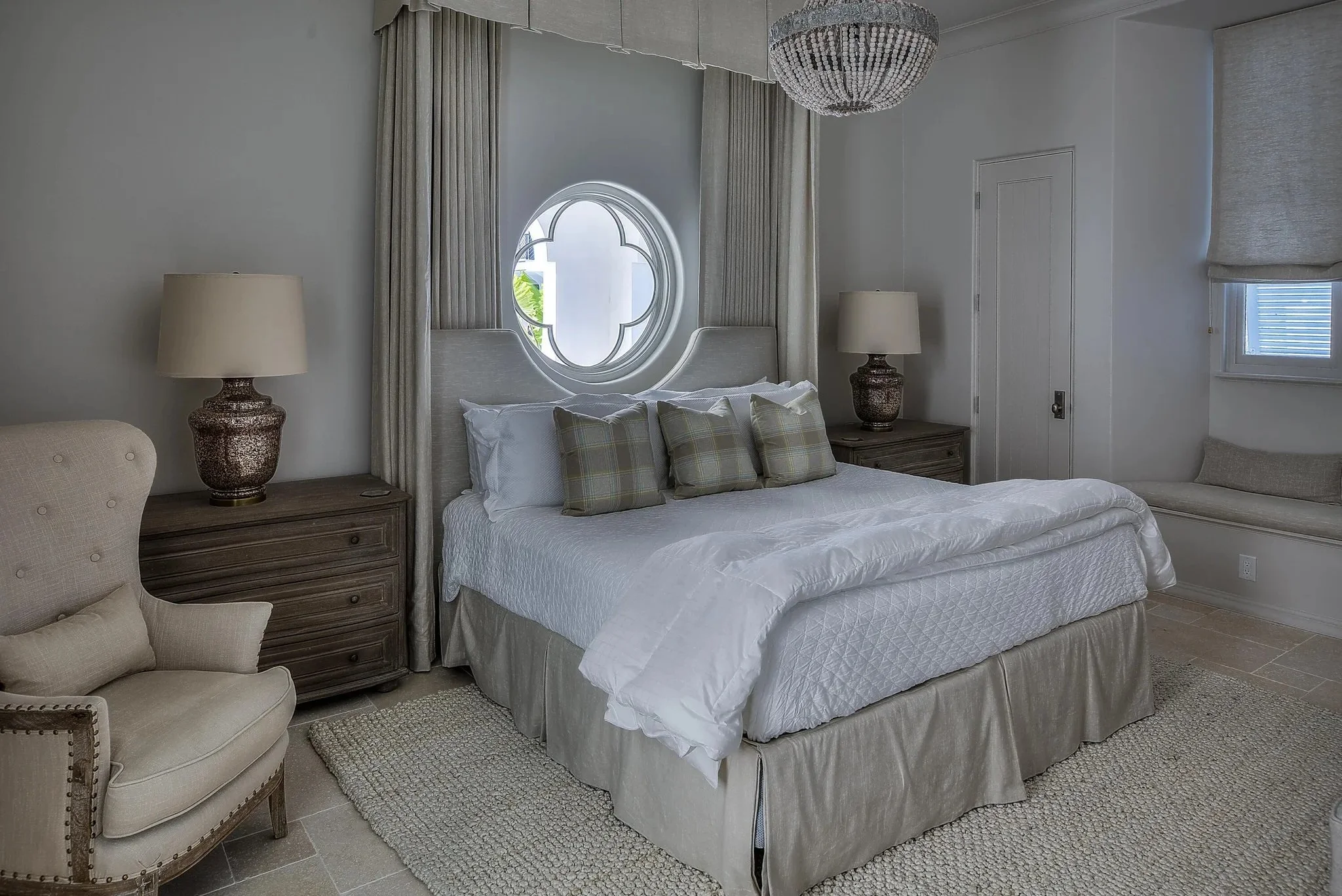 A bedroom with a king-sized bed, white bedding, and four plaid pillows; two matching nightstands with large lamps, an armchair on the left, a window seat with cushion on the right, a circular window behind the bed, and a chandelier hanging from the c