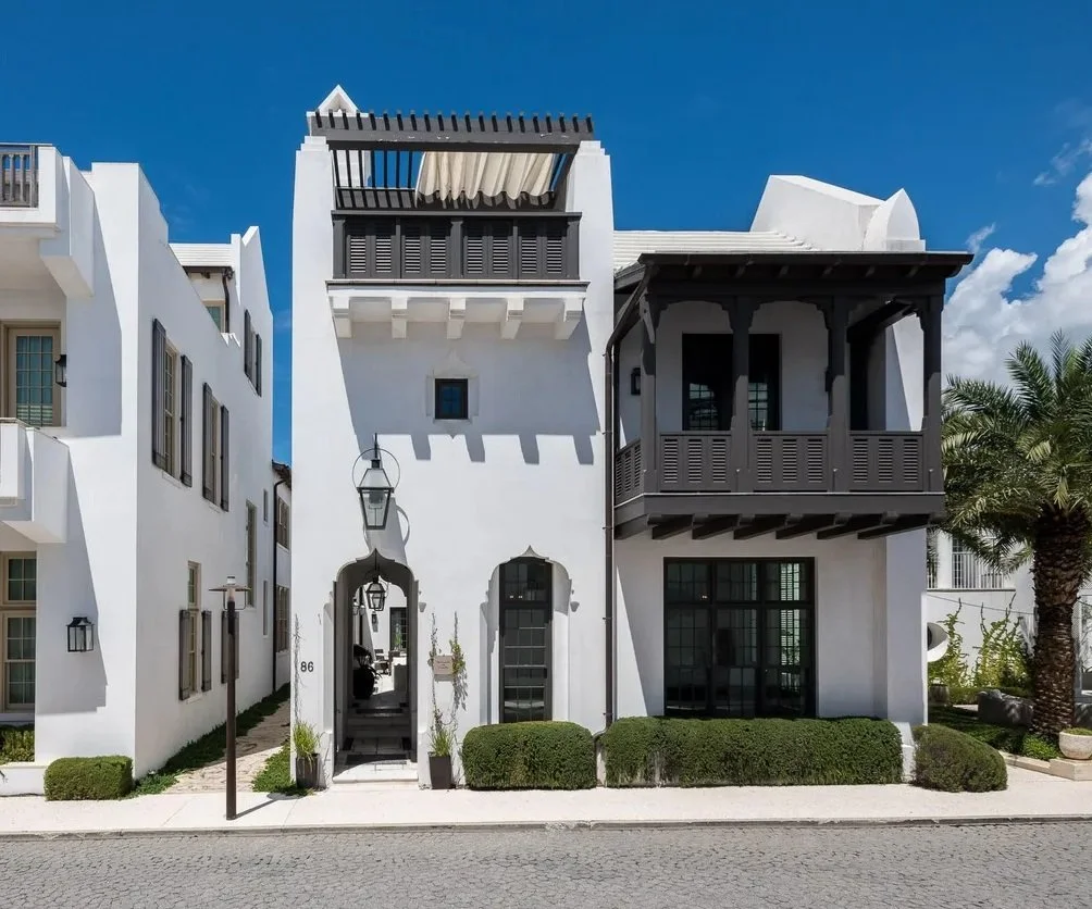 White Mediterranean-style house with black accents, a balcony, and a small front garden with bushes and palm trees