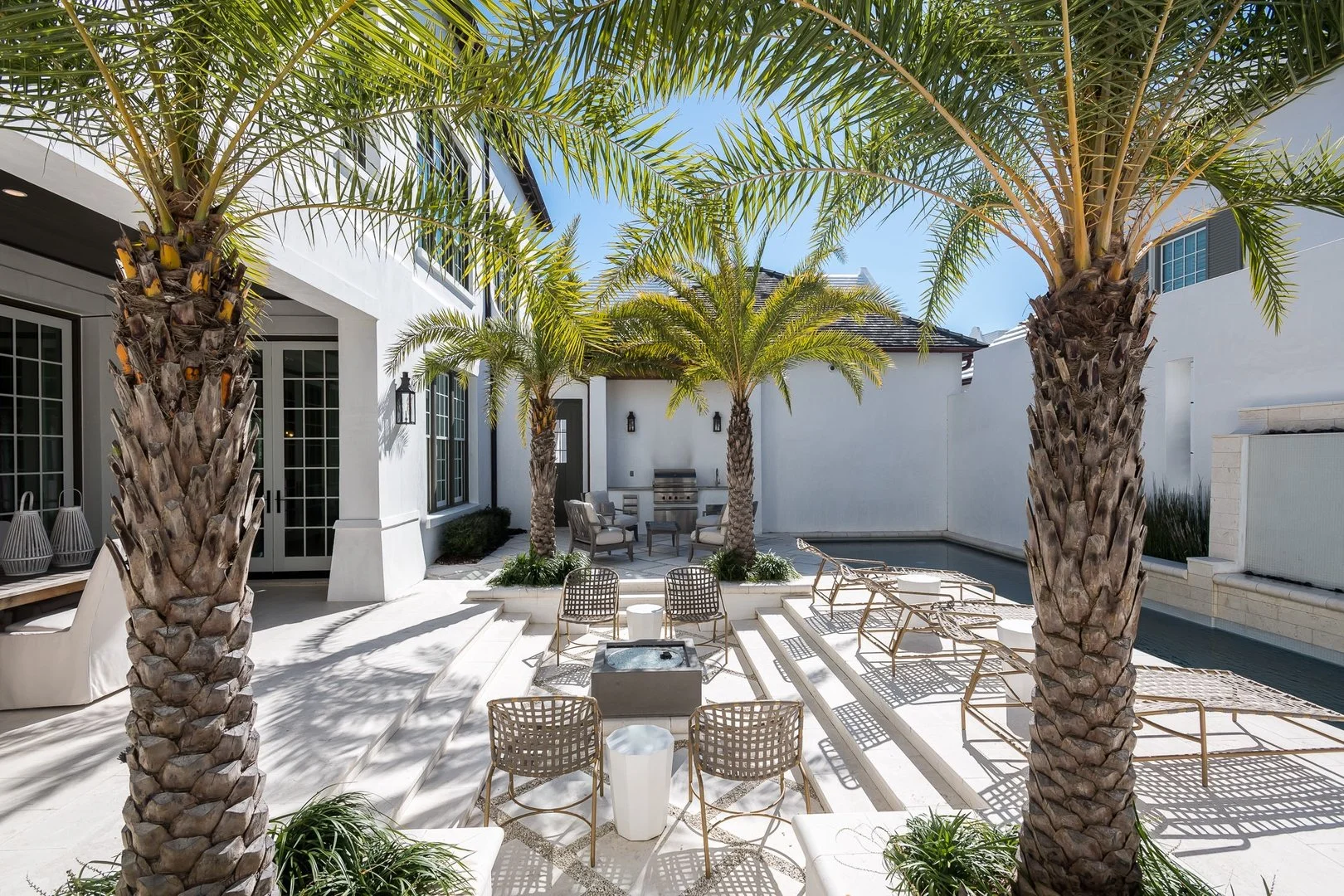 Luxury backyard patio featuring two large palm trees, outdoor seating, a built-in firepit, a rectangular pool with lounge chairs, a barbecue grill area, and modern white walls with black accents under a clear blue sky.