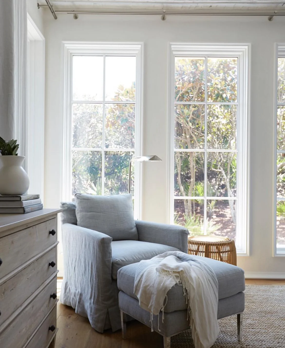 Bright living room with large windows, a gray armchair with a matching ottoman covered with white fabric, a white throw blanket, a wicker side table, a white dresser with black knobs, and a white vase with greenery on top.