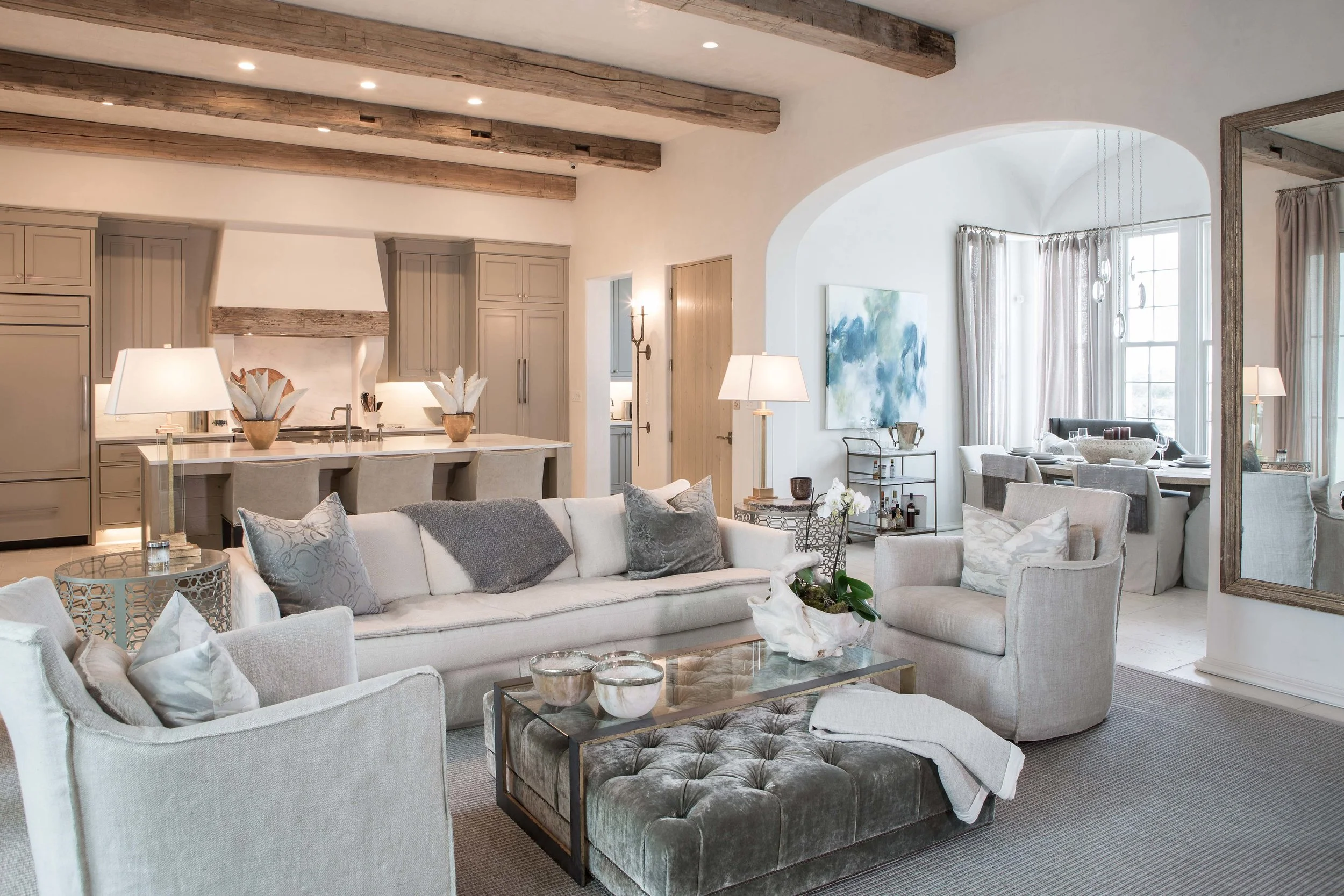 Living room with white sofas, armchairs, and a glass coffee table, open to a dining area with a large window, curtains, and artwork on the wall.