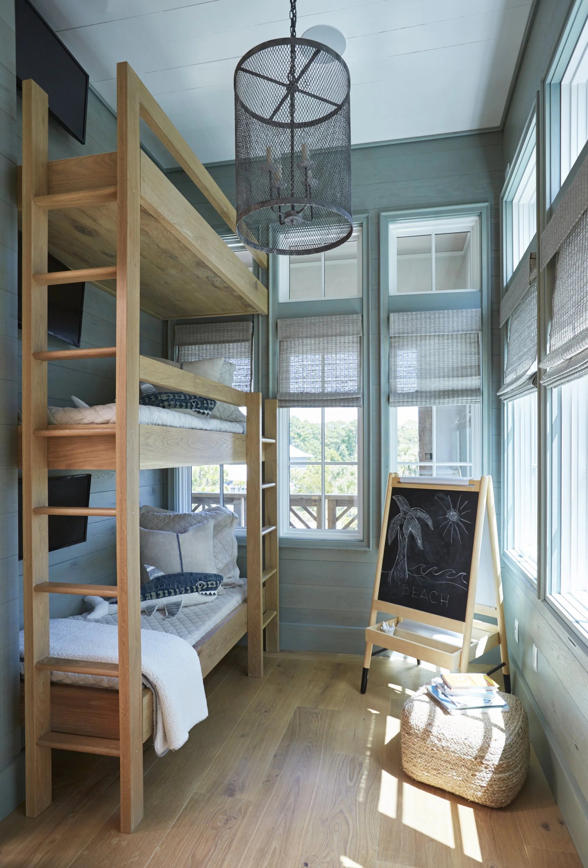 Cozy room with wooden bunk beds, large windows with blinds, a black chalkboard with a beach drawing, and natural sunlight.