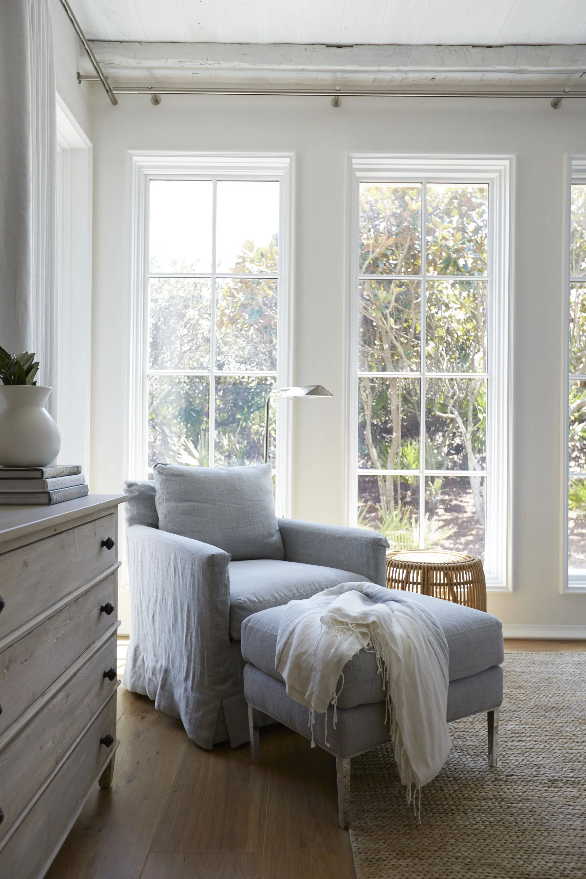 Sunlit living room with large windows, a gray armchair with a matching ottoman, a white throw blanket, a wooden side table, and a dresser with plants and books.